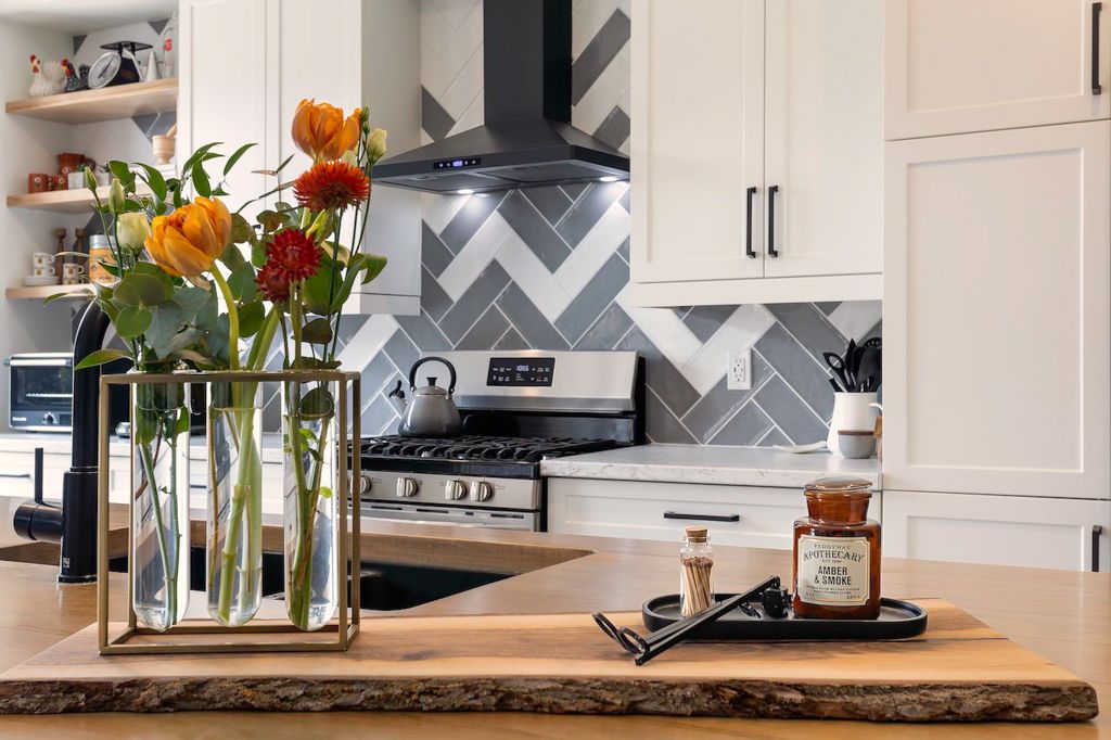 Modern white kitchen with herringbone tile backsplash, stainless steel range, and fresh flowers on a live-edge wood island.