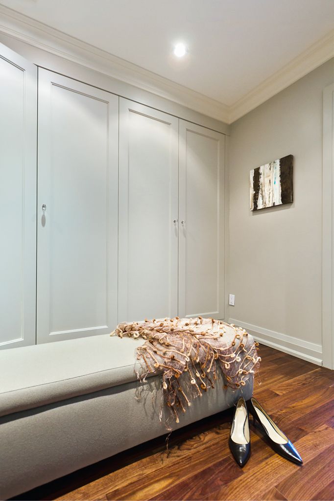 Elegant walk-in closet with built-in white wardrobe doors, hardwood floors, and upholstered bench with decorative throw.