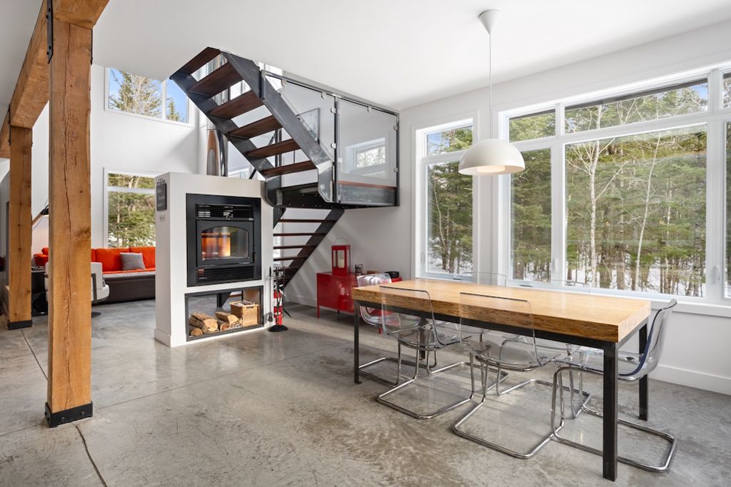 Contemporary open-concept dining area with wooden table, steel staircase, fireplace, and floor-to-ceiling windows overlooking snowy trees.