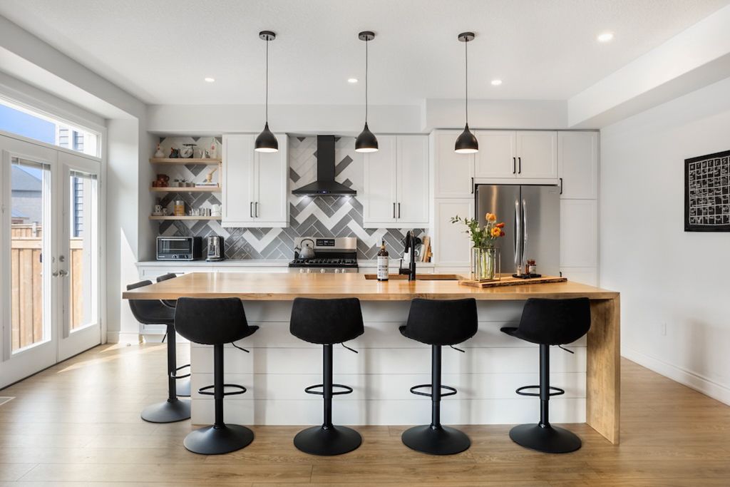 Modern white kitchen with large island, butcher block countertop, black barstools, and pendant lights.