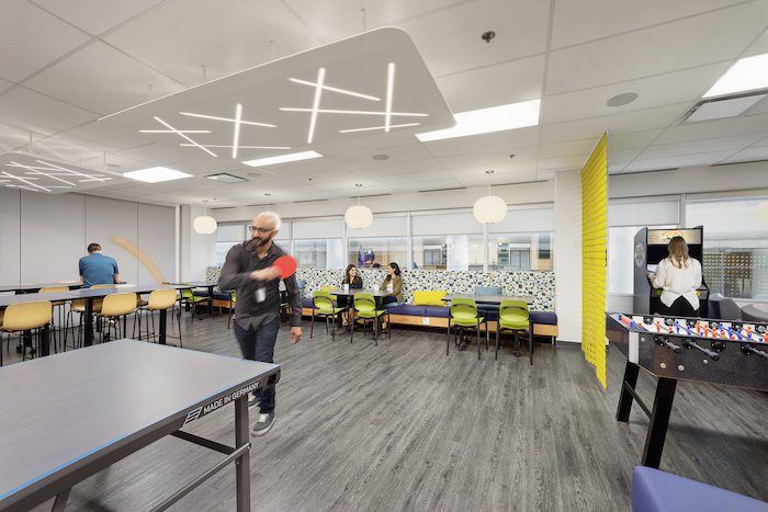 Bright breakroom with yellow accents, foosball table, pendant lights, and employees relaxing at cafe tables.