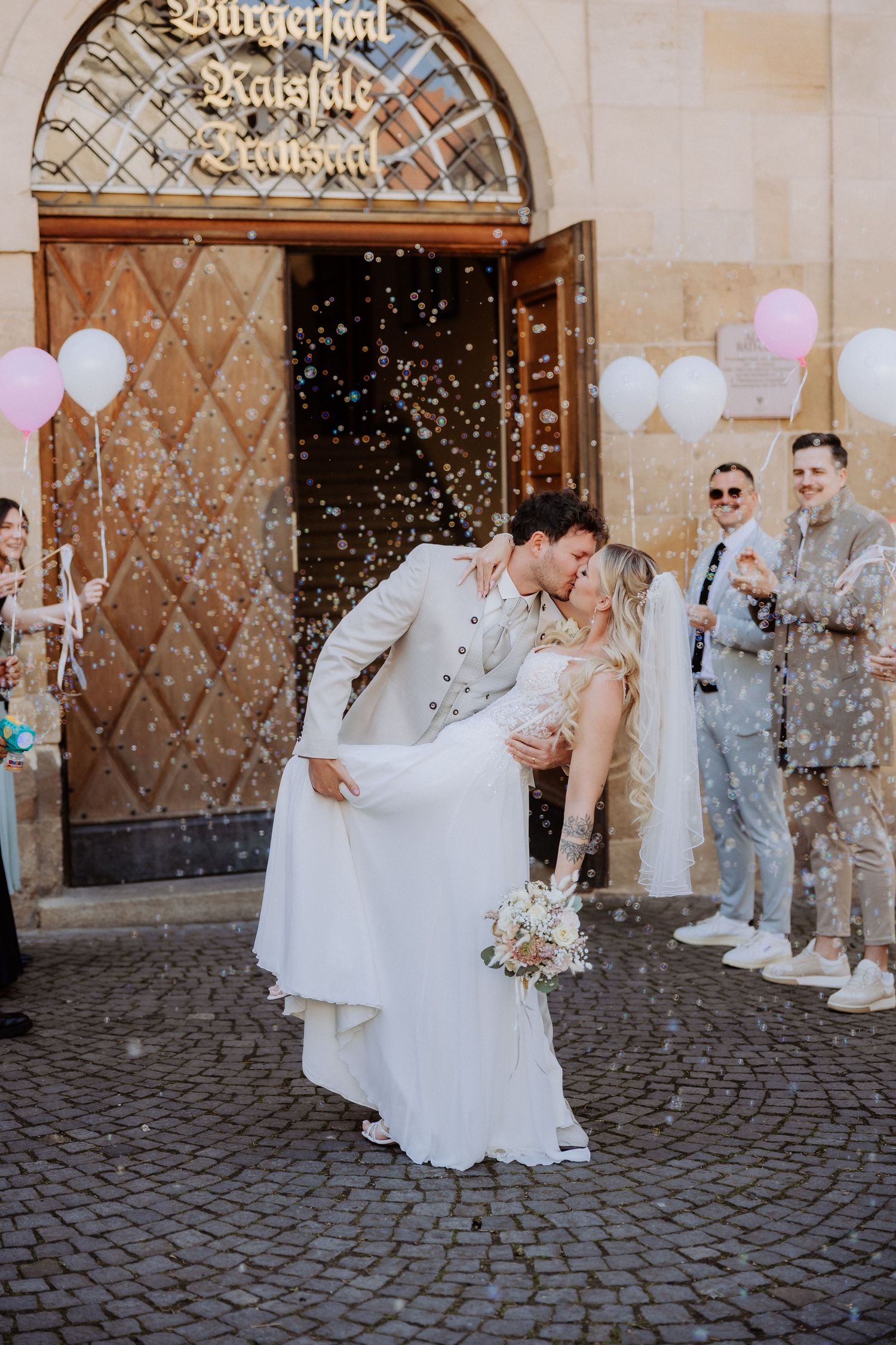 Wedding couple celebrates with confetti and bubbles on cobblestone street while guests look on with joy.