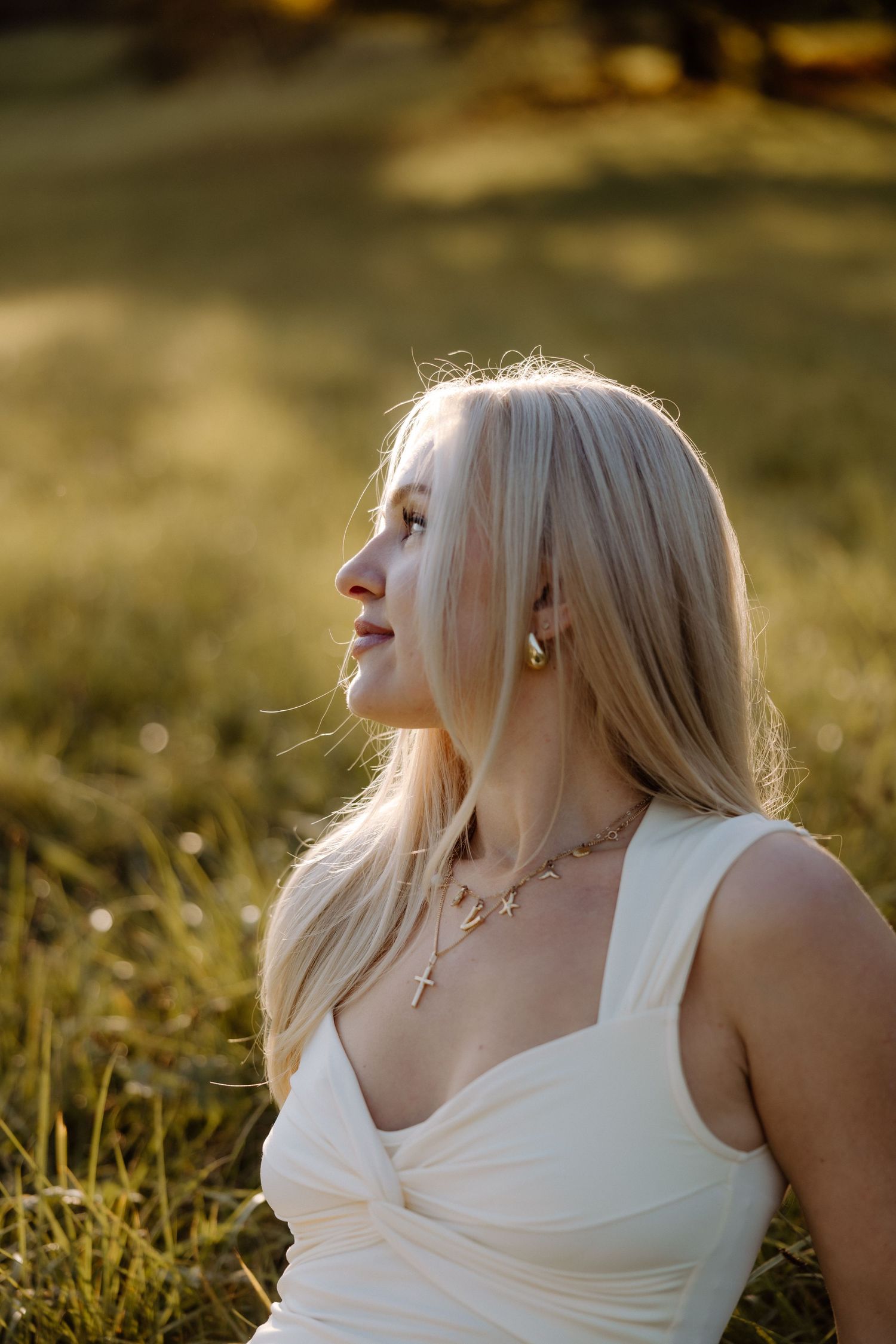 Person with platinum blonde hair in white tank top poses in golden sunlight against natural outdoor setting.