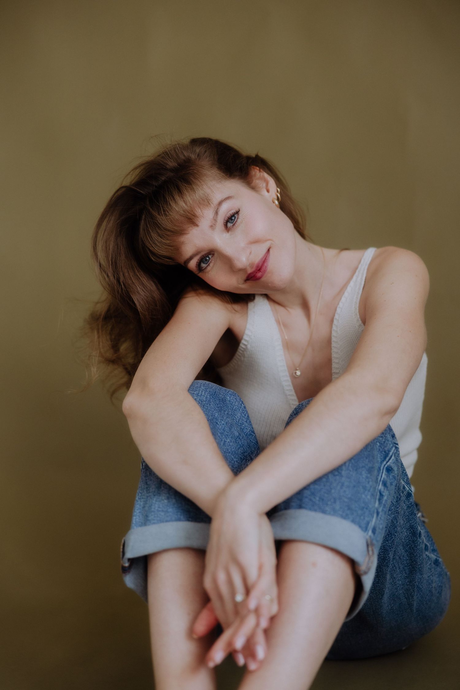 A person in denim poses against a neutral background while resting their head on their knee.