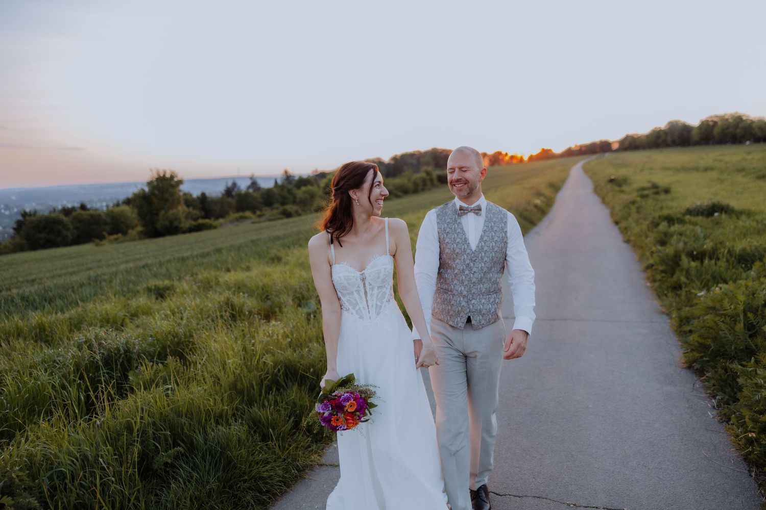 Bride and groom walking hand in hand along a scenic countryside path at sunset, bride holding a colorful bouquet.