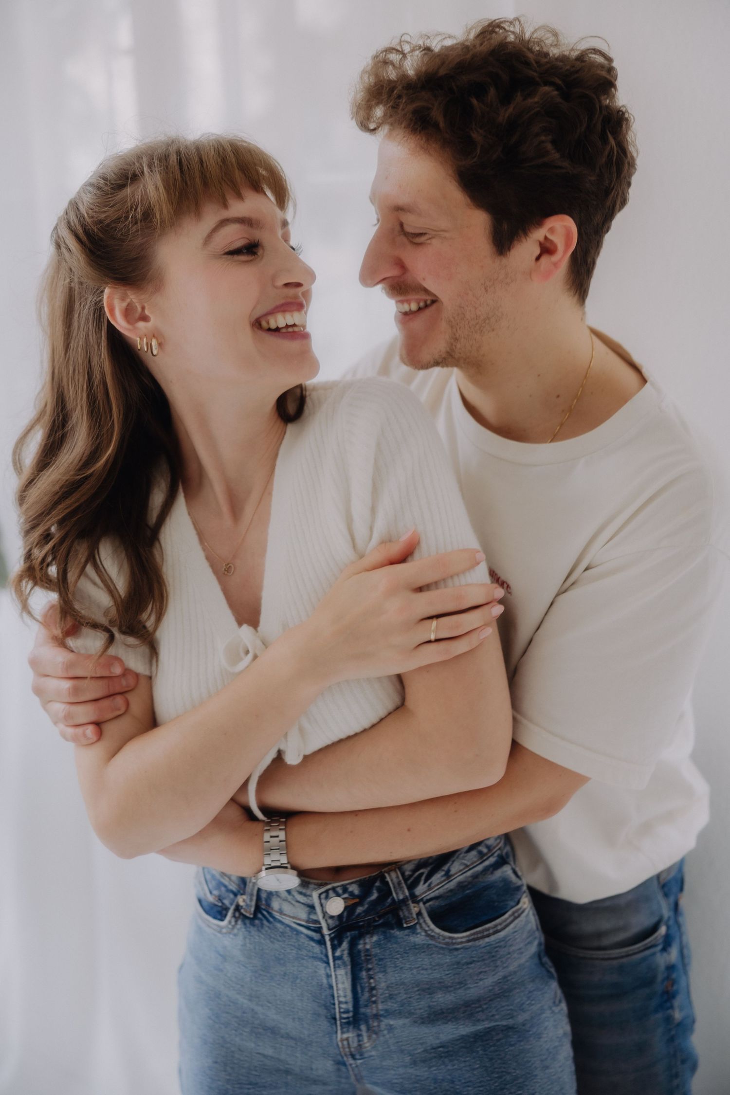 A romantic couple in casual white sweaters and jeans embraces and laughs together against a light background.