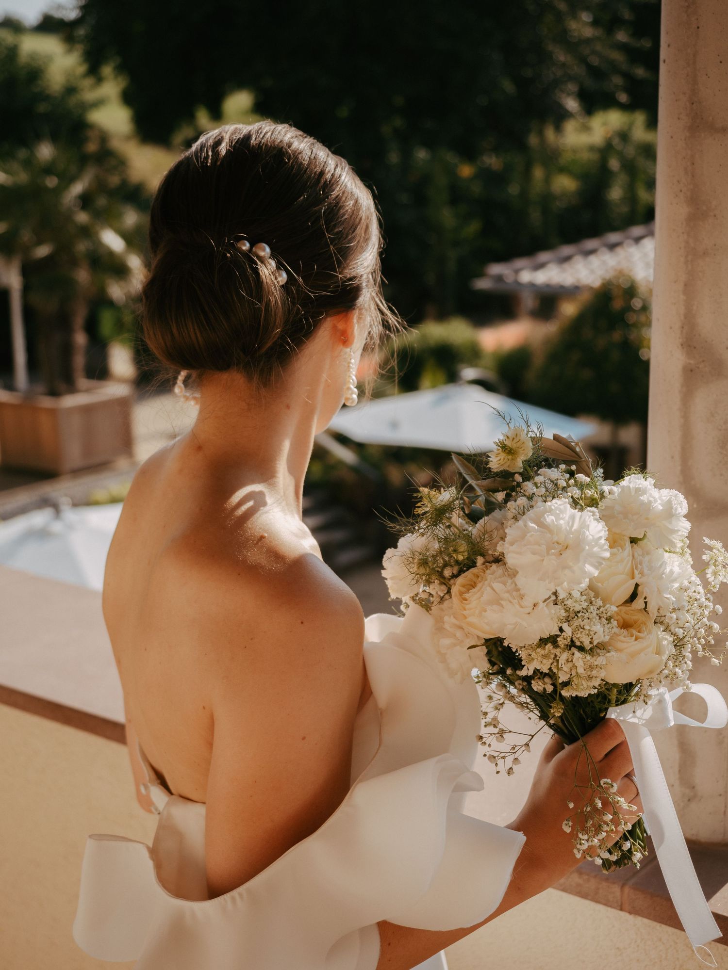 Back view of a bride's elegant white dress and floral bouquet against a bright outdoor setting.
