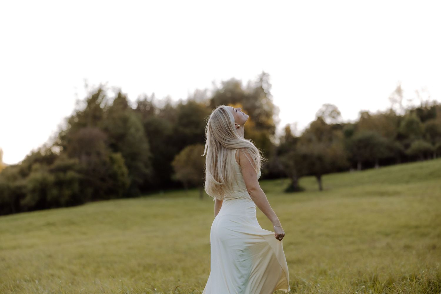 A person in a white dress walks through a serene green meadow at sunset with trees in the background.