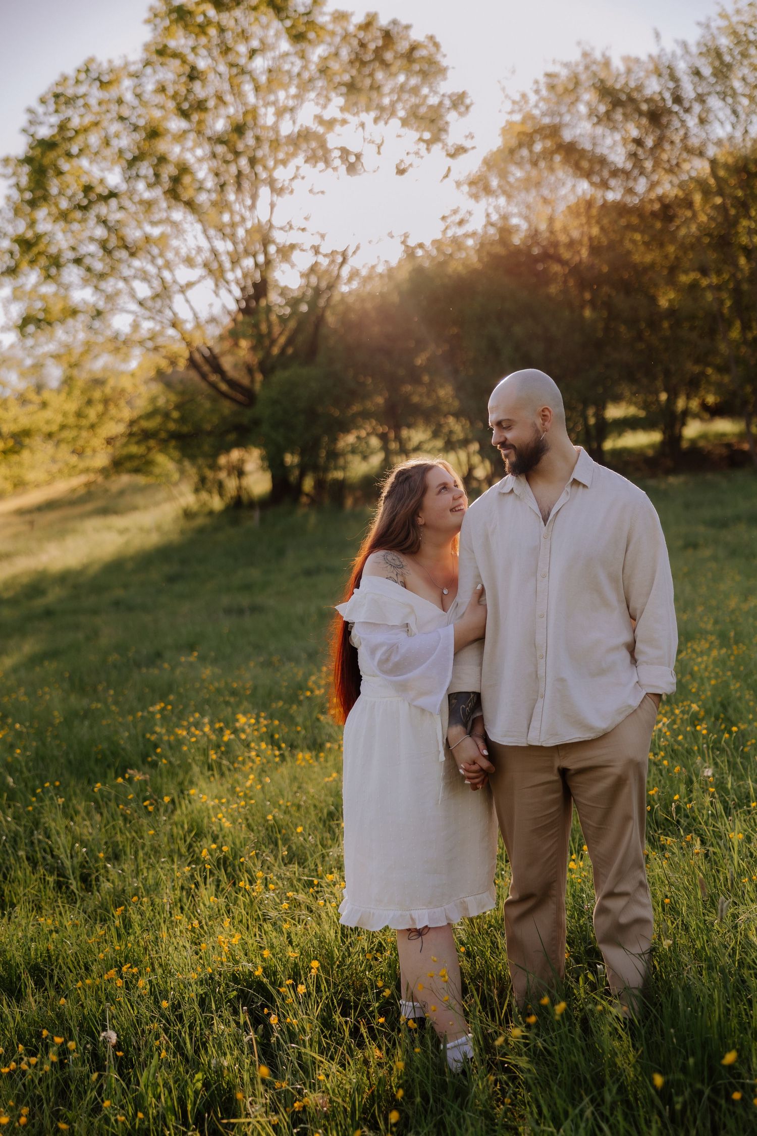 Couple embracing in a sunlit meadow with wildflowers, woman in white dress and man in linen shirt, golden hour.