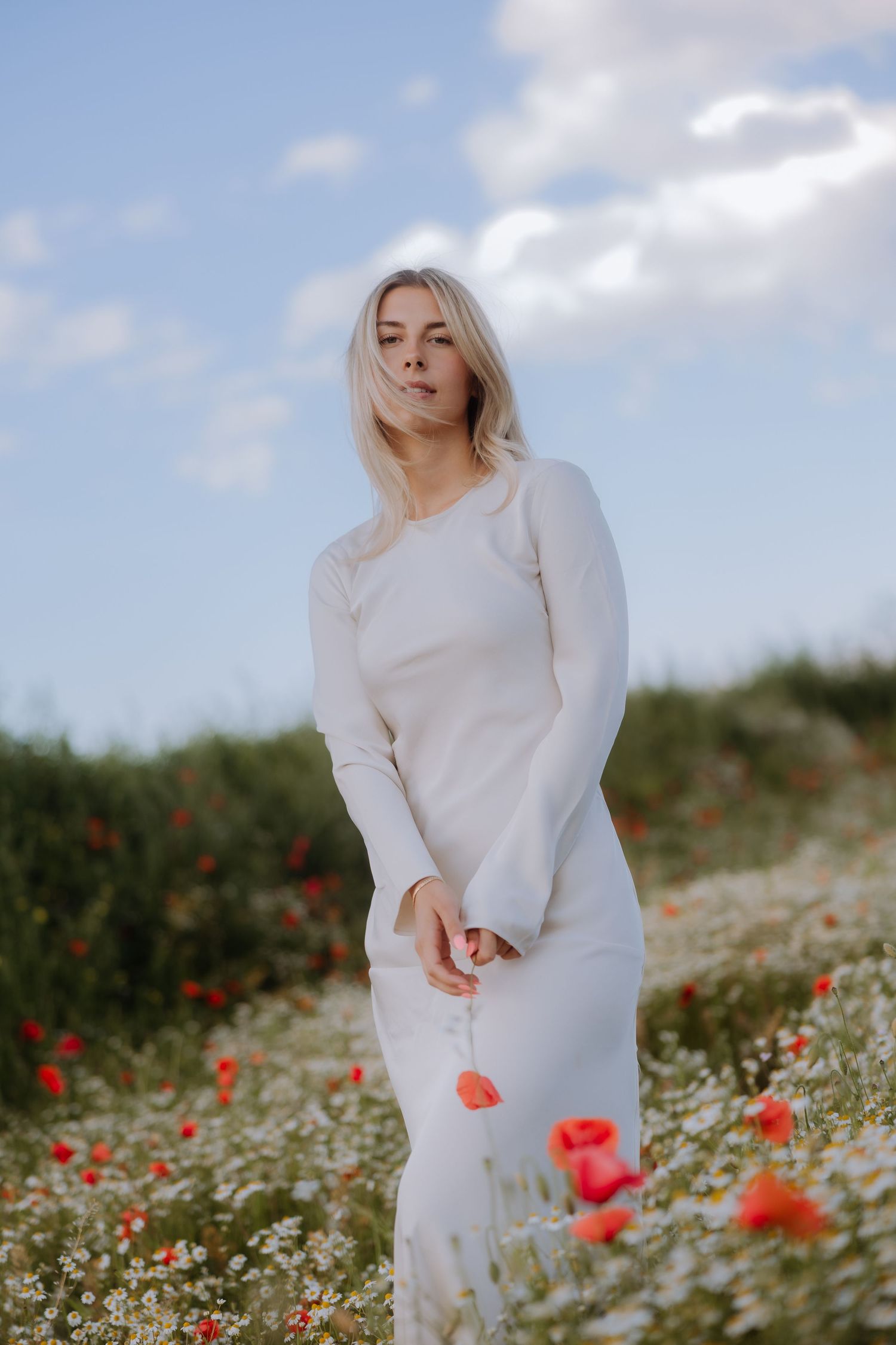 Blonde woman in white dress standing in a field of red poppies and daisies under a blue sky.