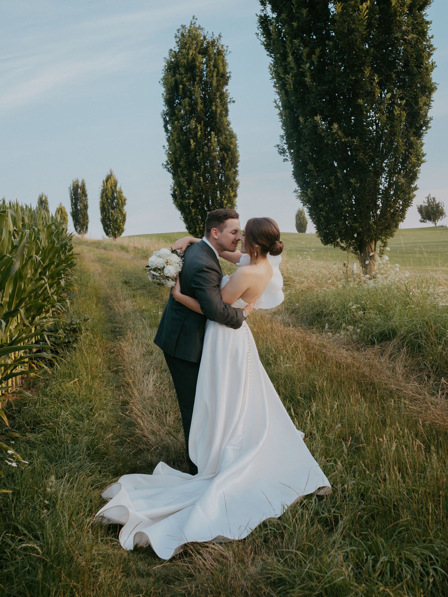 Newlyweds dance and twirl in an outdoor field with tall poplar trees in the background.
