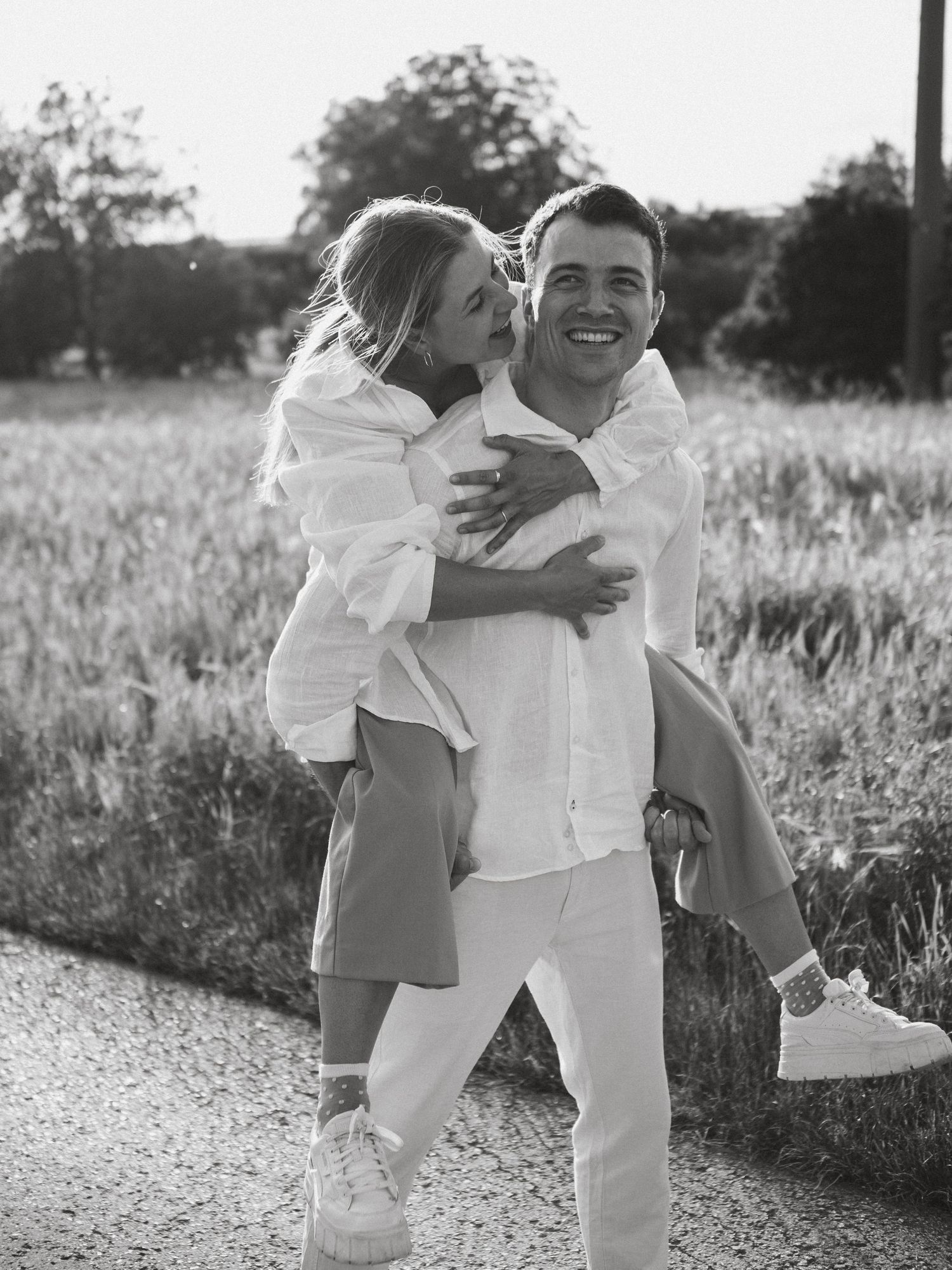 A series of black and white photos showing a couple in white outfits embracing playfully in a sunlit field.
