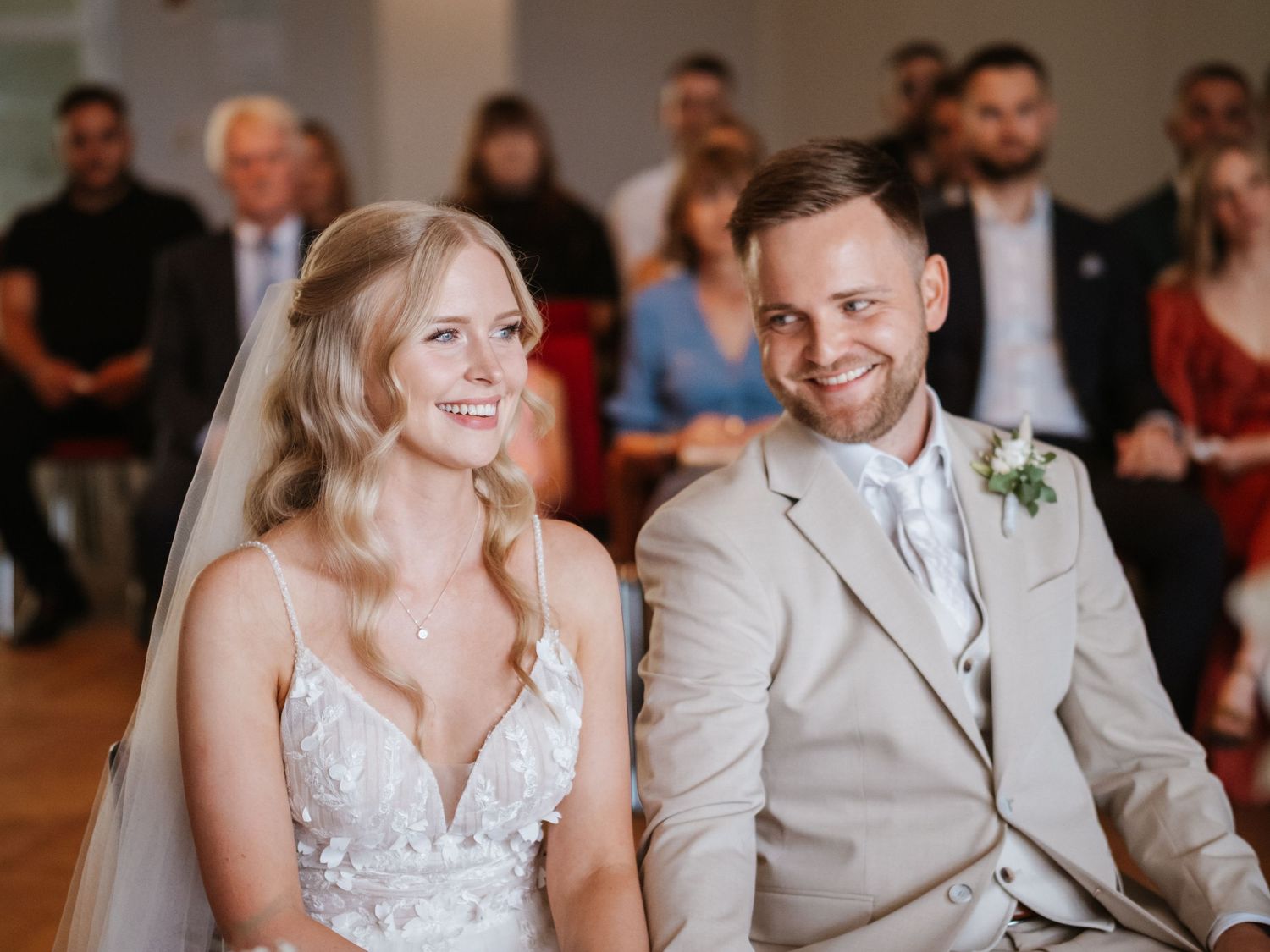 A happy couple smiles together during their wedding ceremony, with the bride in white lace and groom in tan suit.