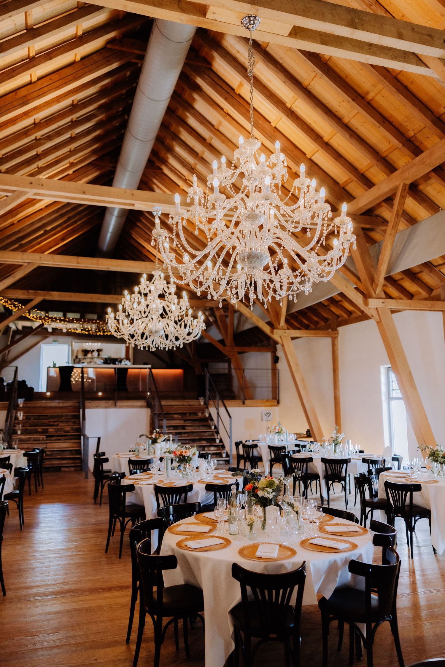 Elegant barn wedding venue with crystal chandeliers, round tables set with white linens, and exposed wooden beam ceiling.