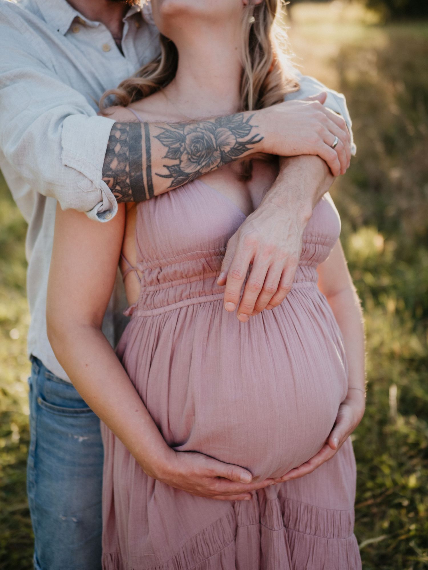 A couple embraces in a sunlit field while cradling a pregnant belly in a dusty pink maternity dress.