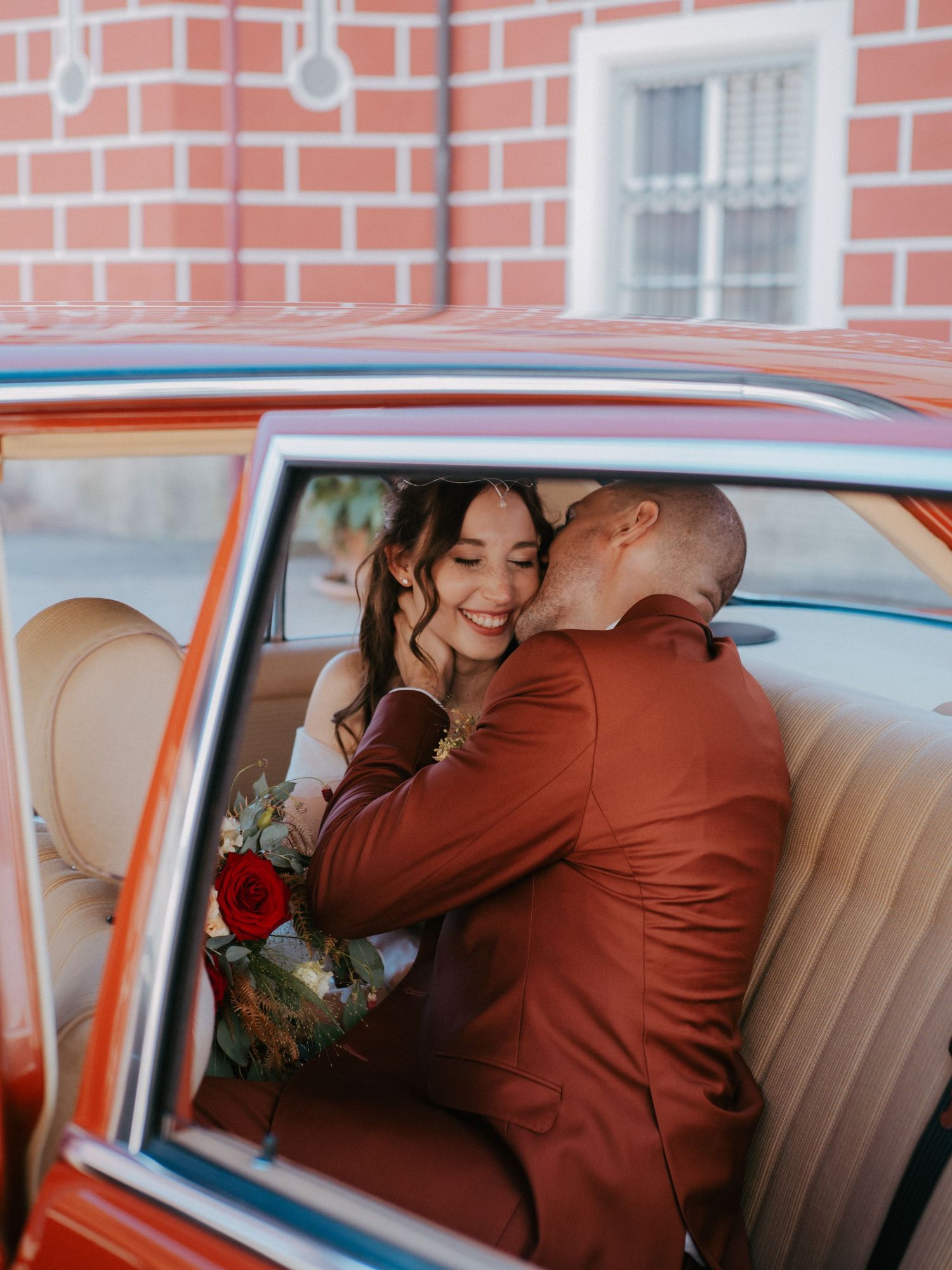 Romantic wedding couple shares intimate moments in vintage car backseat with brick wall backdrop.