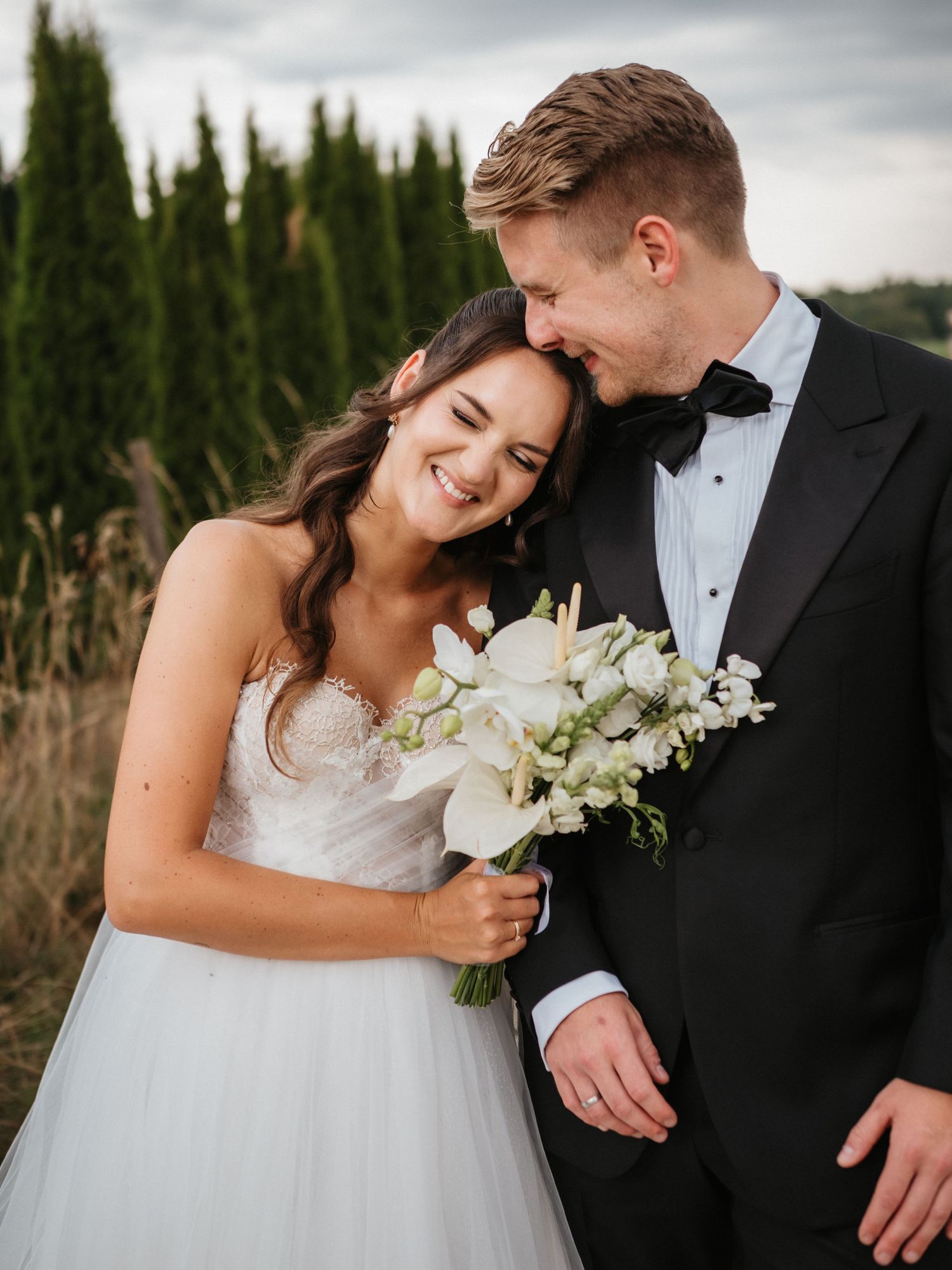 A romantic wedding couple embraces outdoors with cypress trees in background as bride holds white floral bouquet.