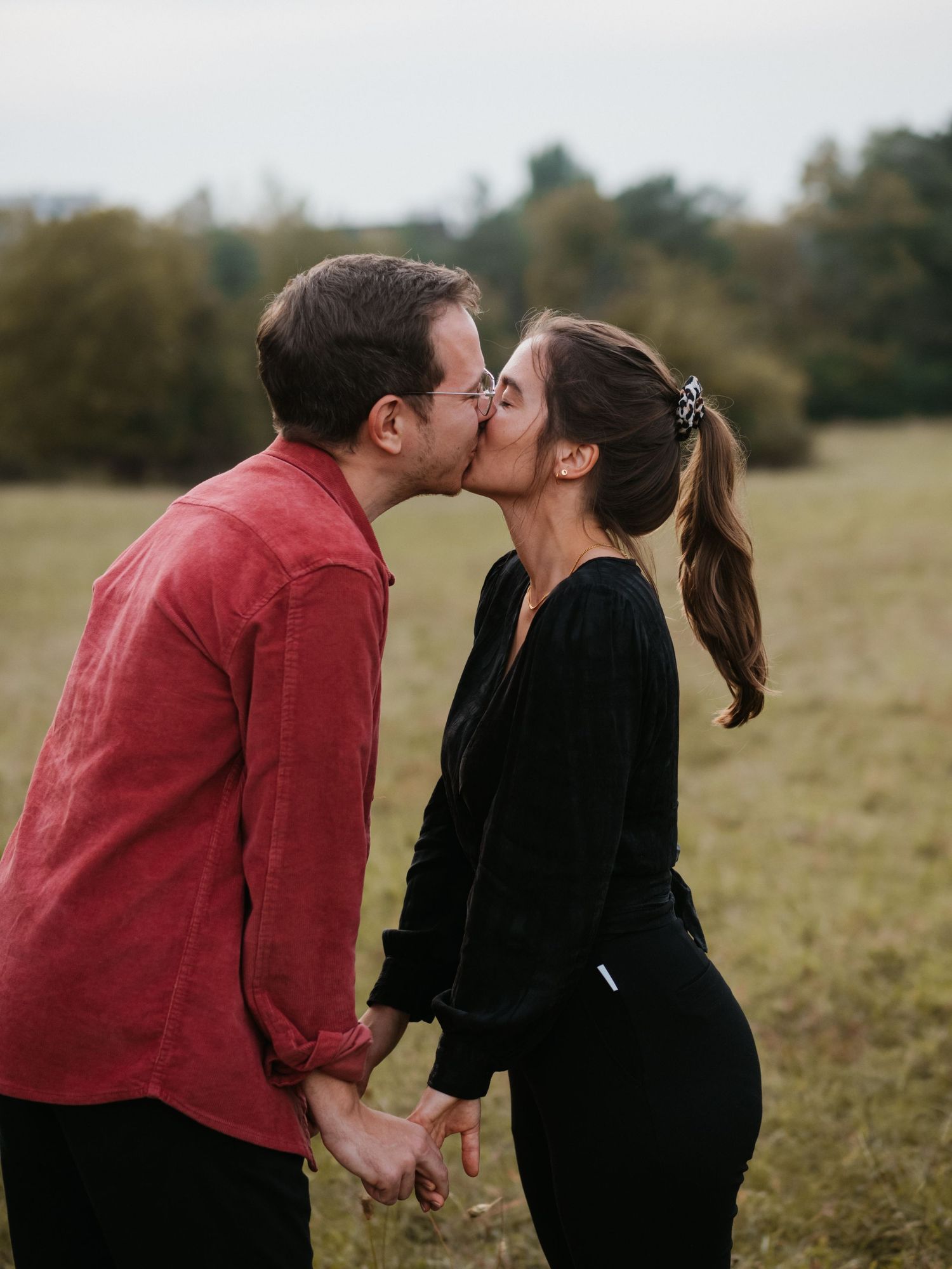 A couple in red and black outfits share a tender kiss in a sunlit field during an outdoor photo session.