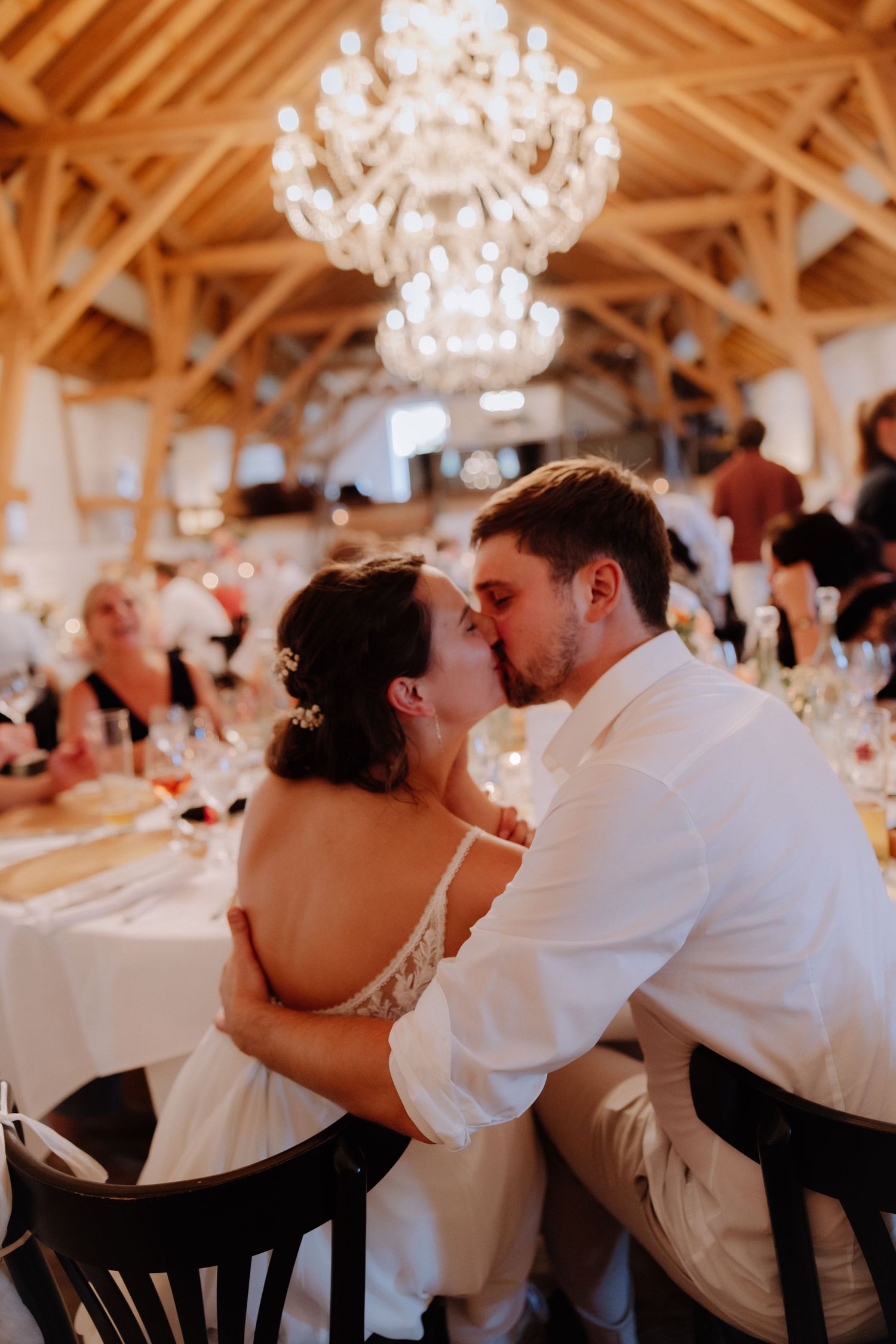 Bride and groom share a romantic kiss at their wedding reception under a crystal chandelier in a rustic barn venue.