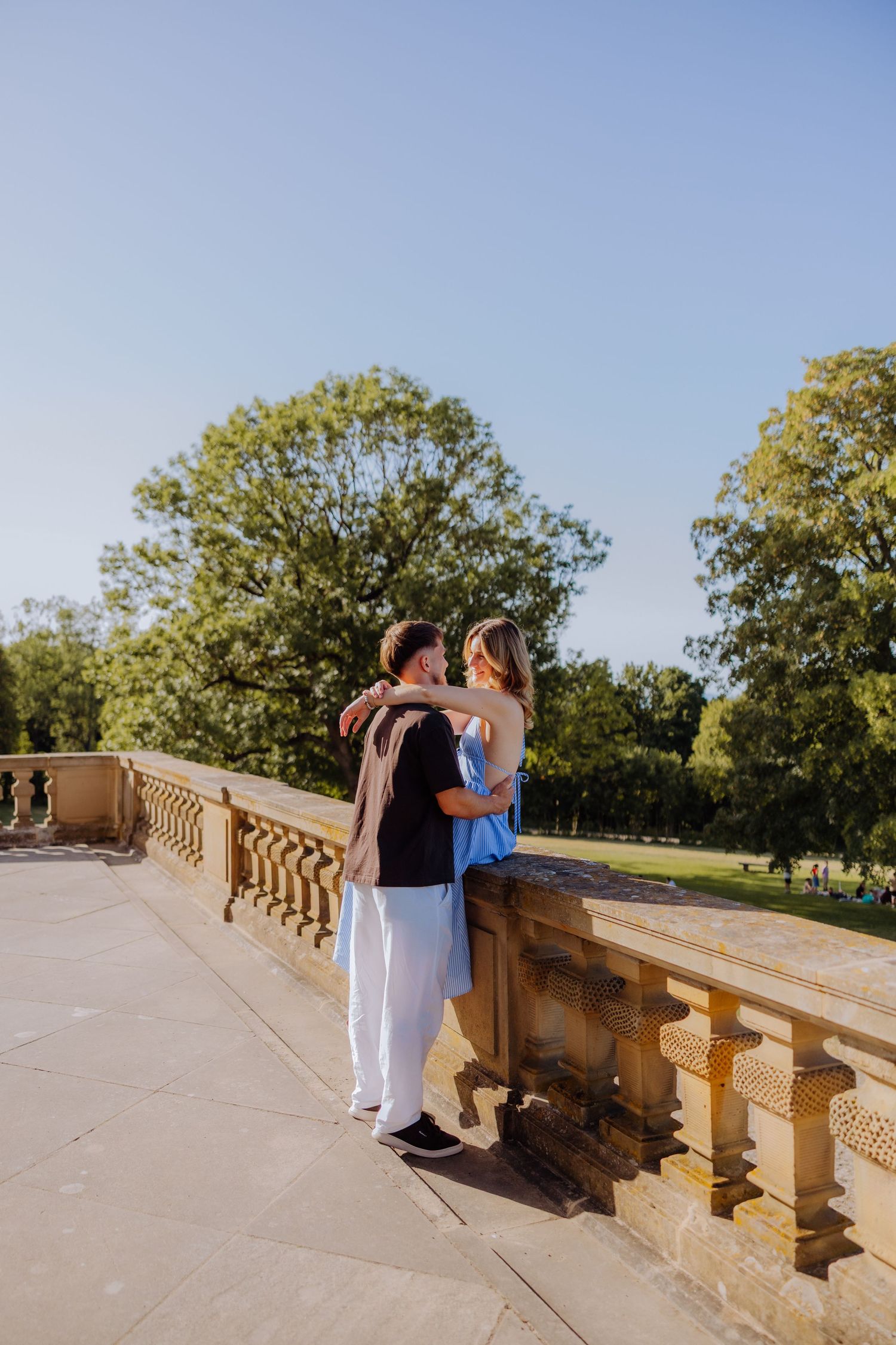 Couple embracing on a stone balcony terrace surrounded by lush green trees under a clear blue sky.