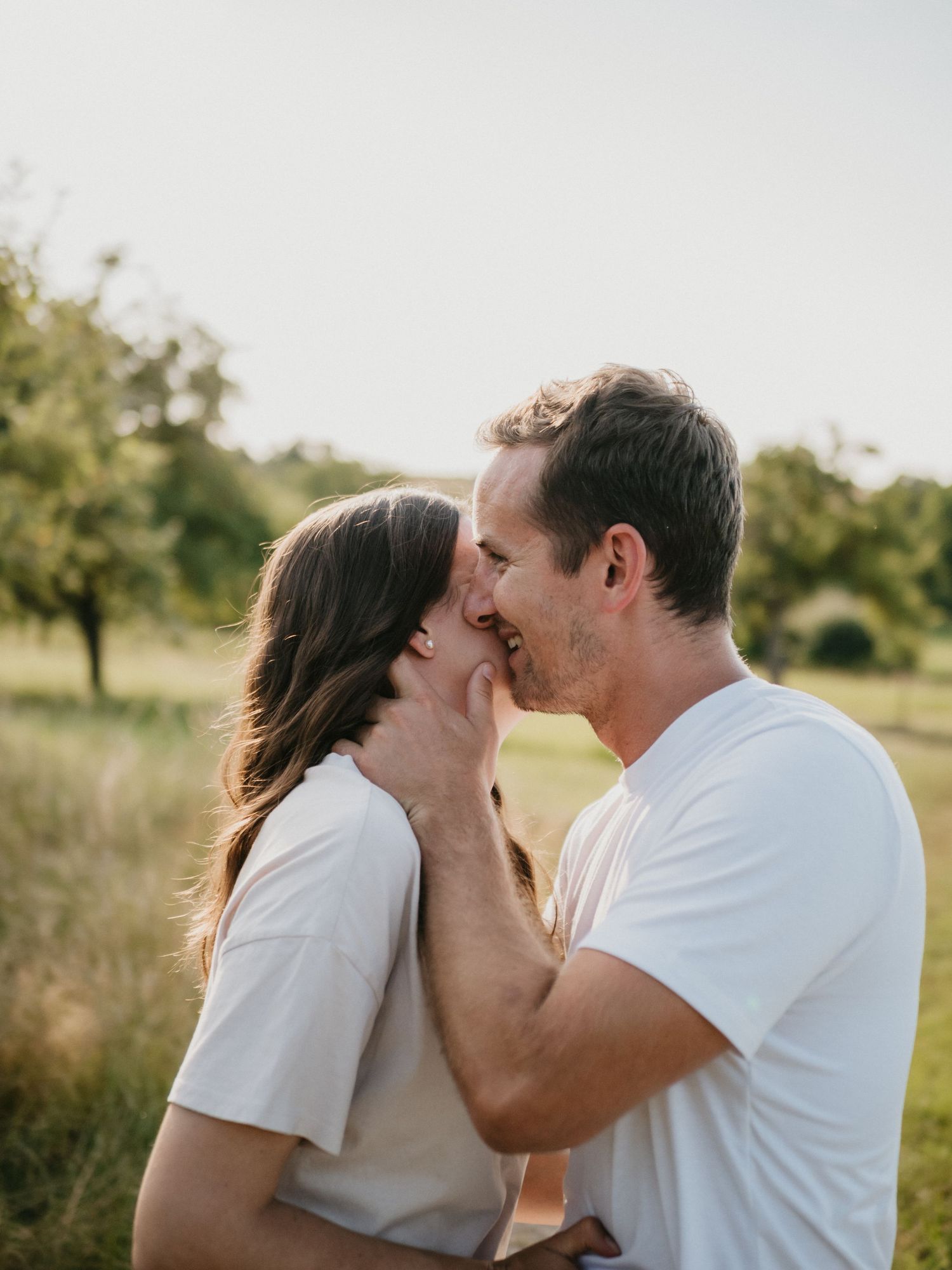 A couple in white shirts share a tender kiss outdoors in a natural setting during golden hour.