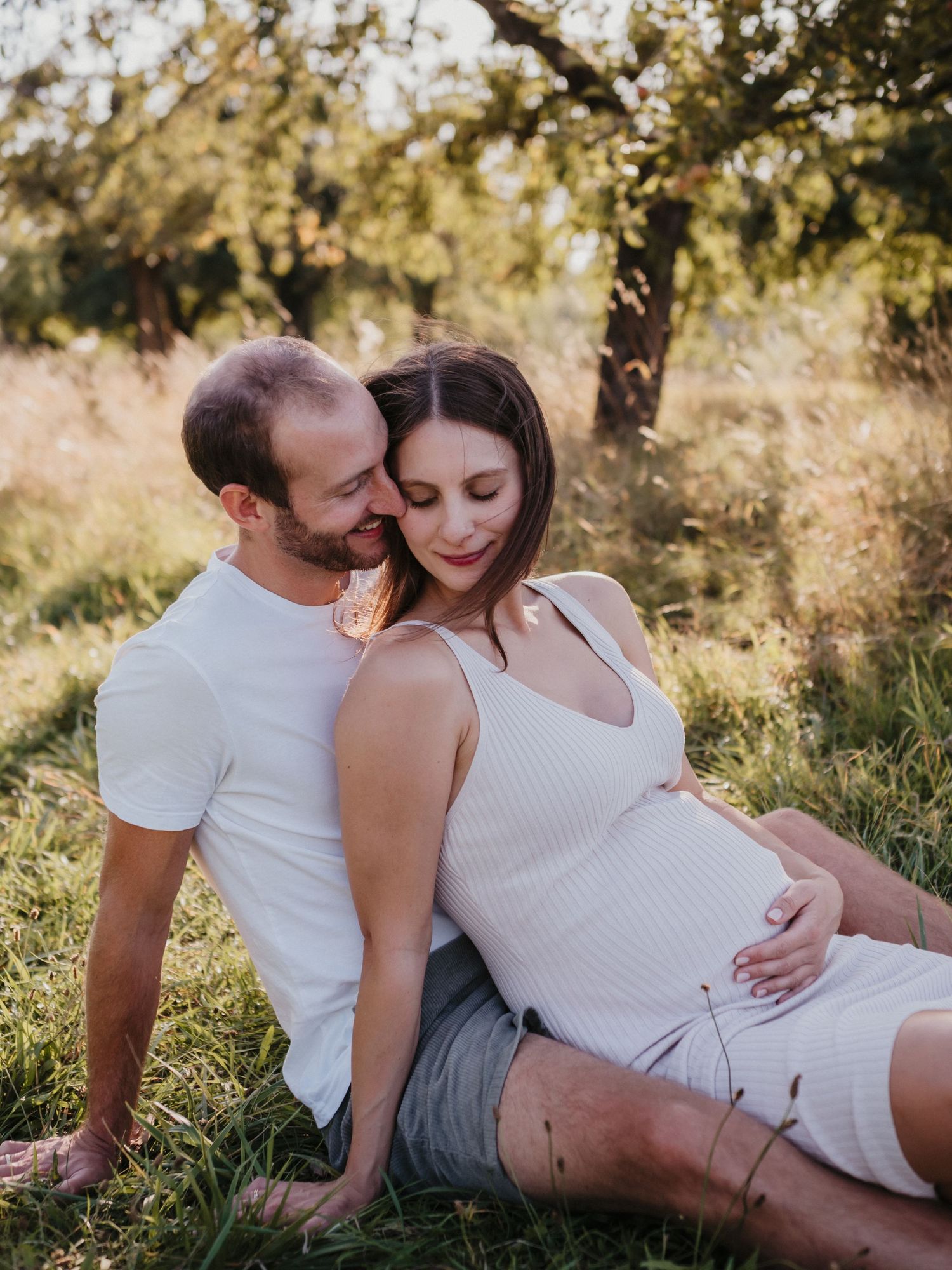 An intimate maternity portrait of an expecting couple sitting together outdoors in warm sunlit nature.