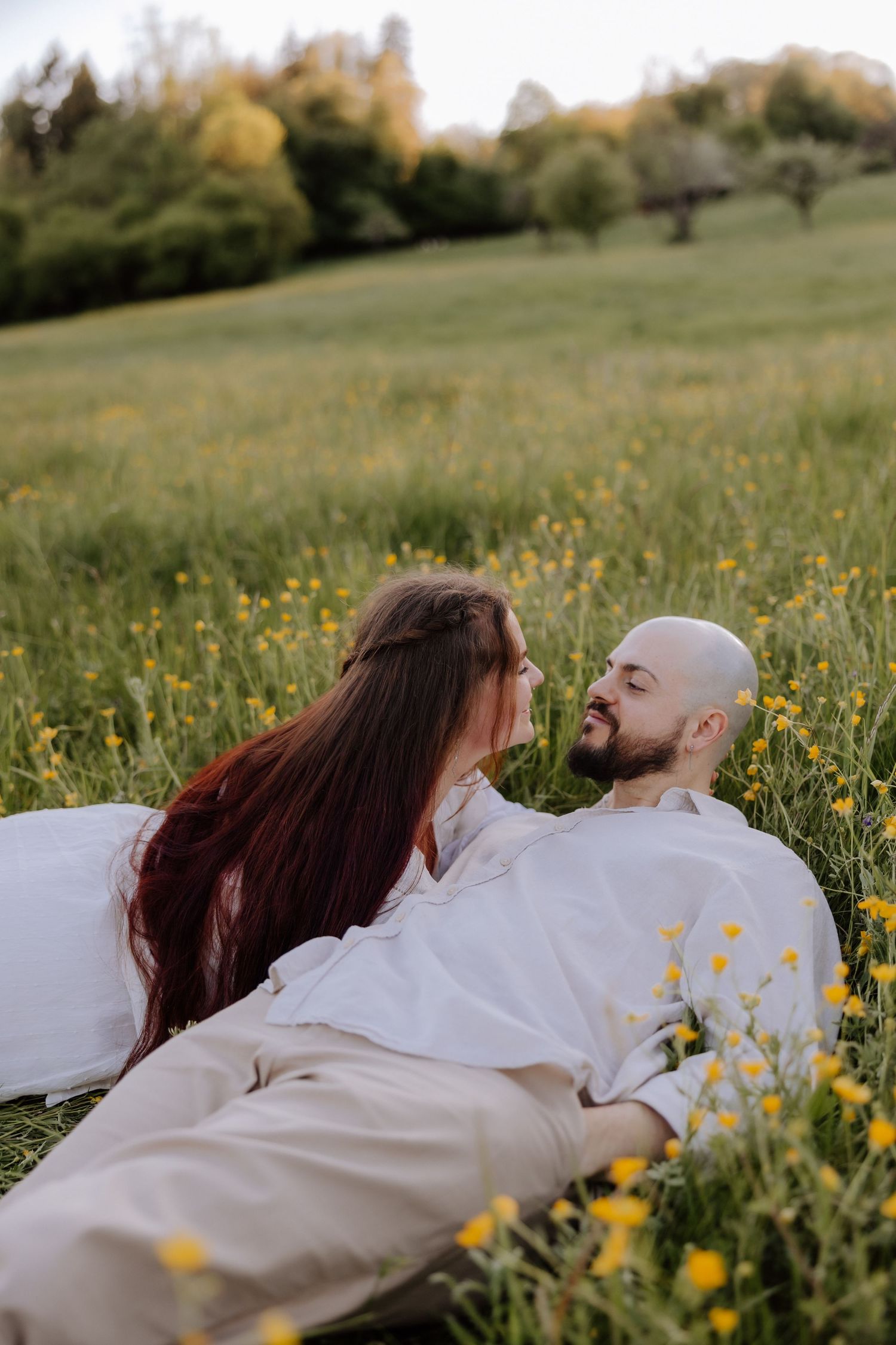 Romantic couple in white outfits lying together in a green meadow with yellow wildflowers, sharing an intimate moment.
