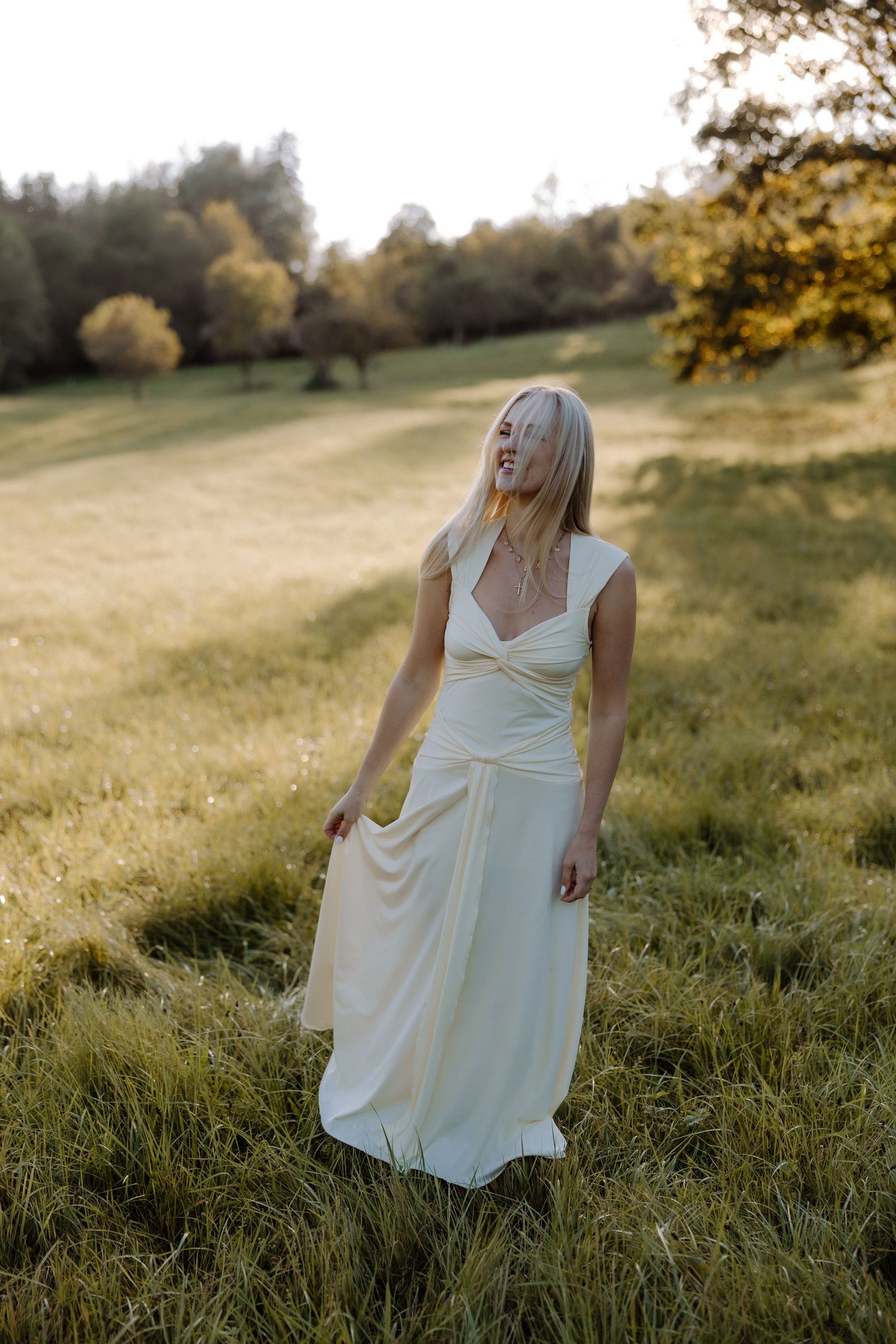 A person in a flowing white dress stands in a sunlit meadow during golden hour with autumn trees in the background.