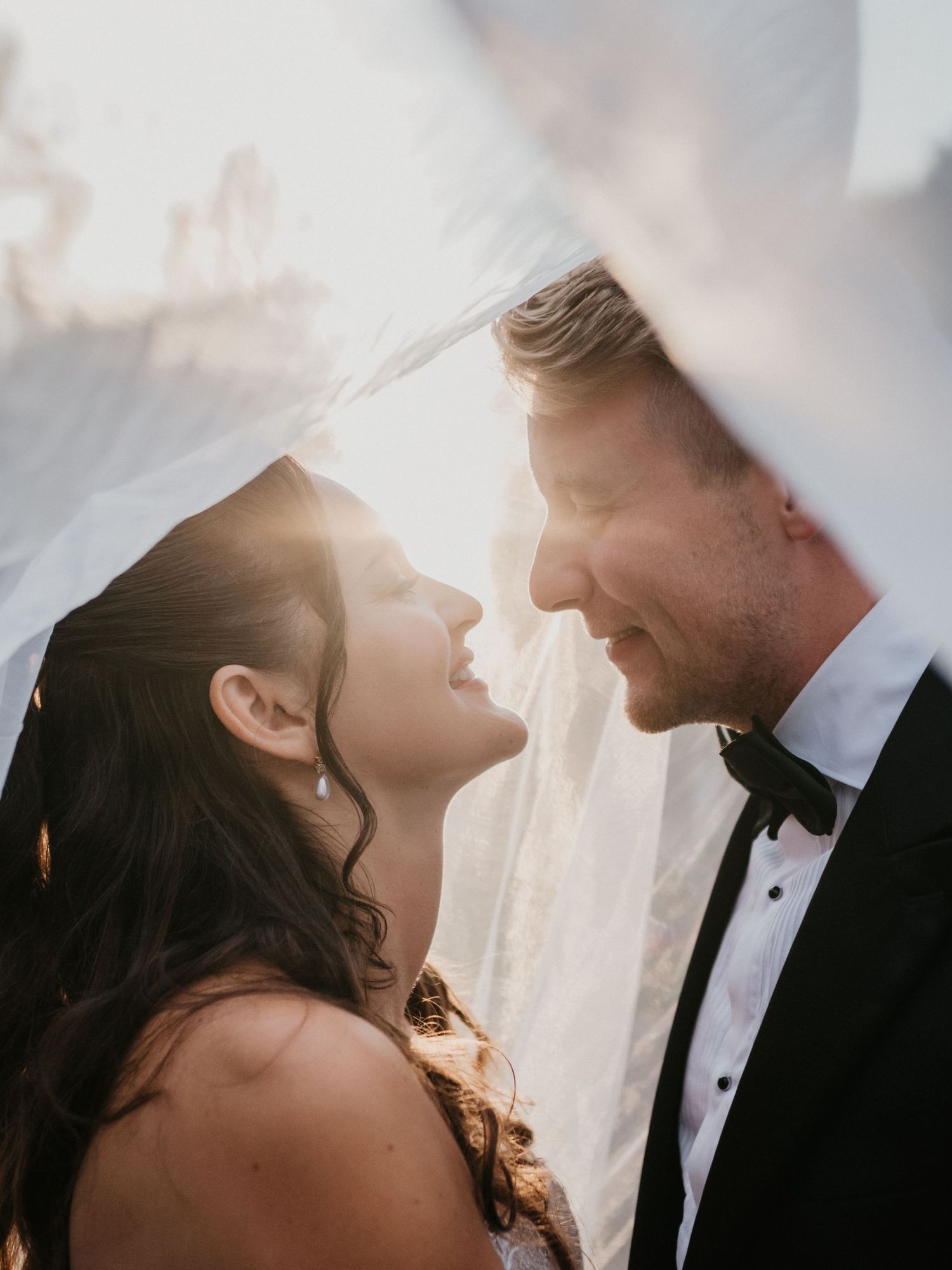 Romantic close-up shot of a couple under flowing white veil with sunlight peeking through.