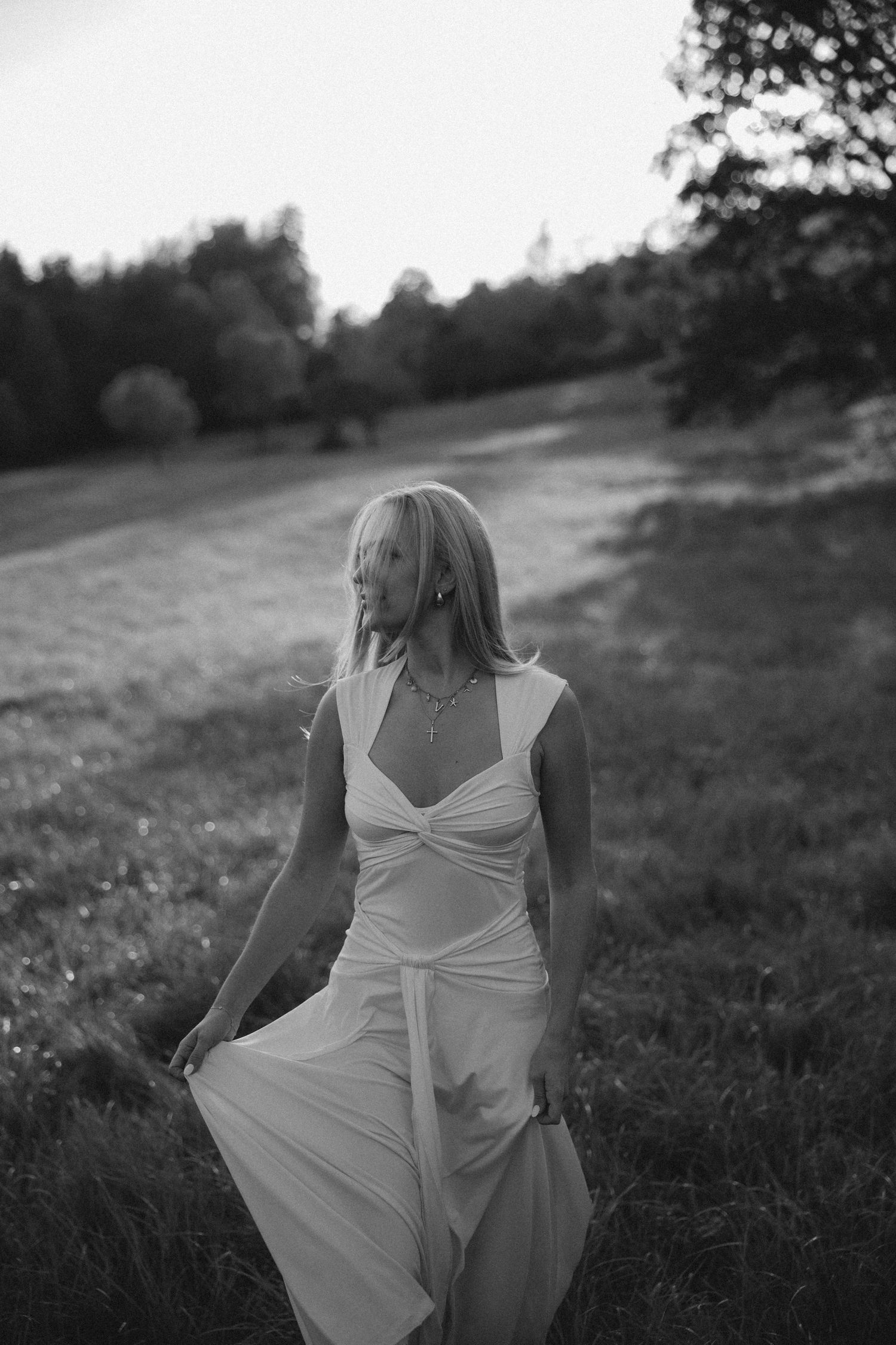 A black and white artistic photograph of someone in a flowing white dress sitting in a grassy meadow at dusk.