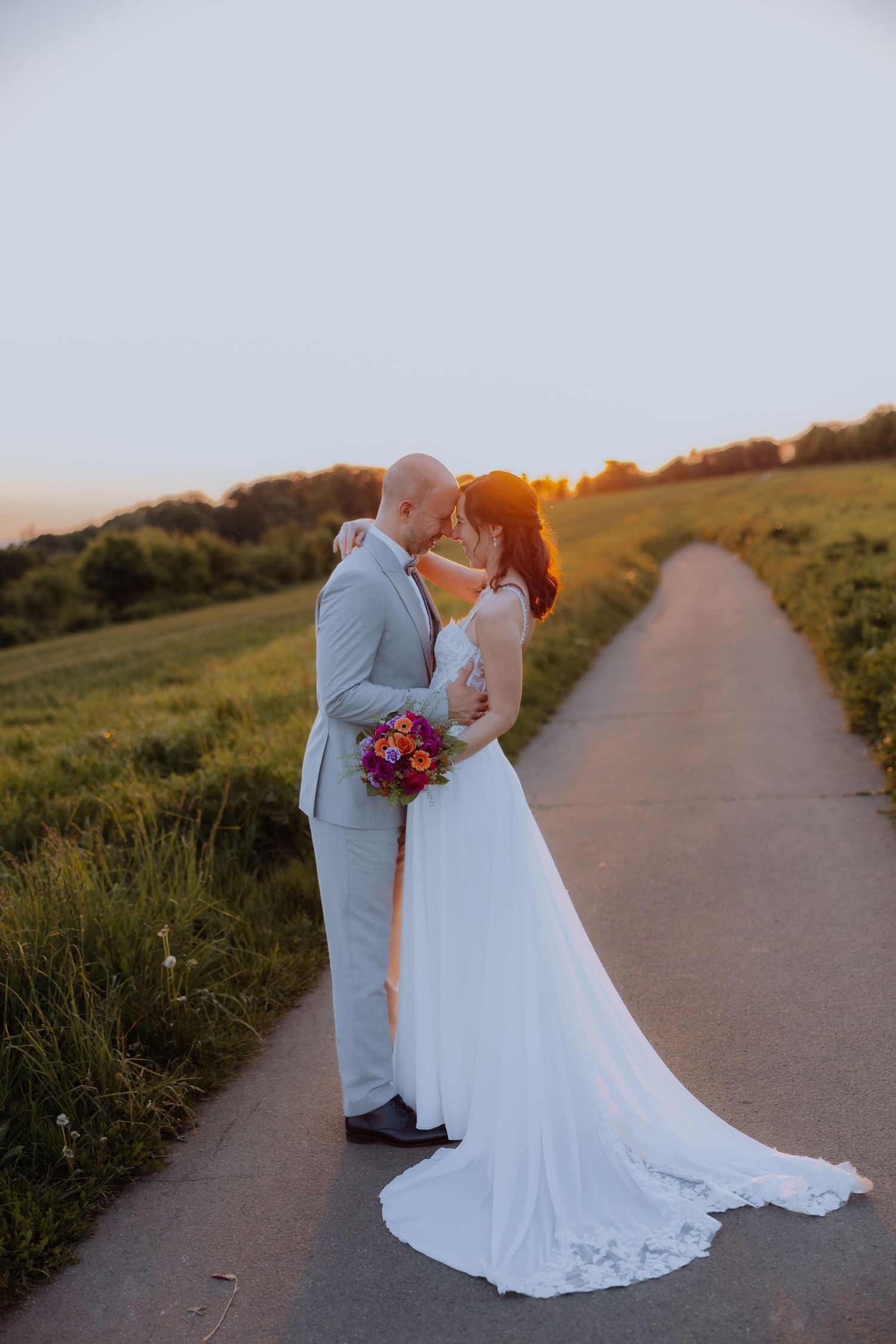 Bride and groom sharing a romantic sunset kiss on a country road, bride holding colorful bouquet in white gown.