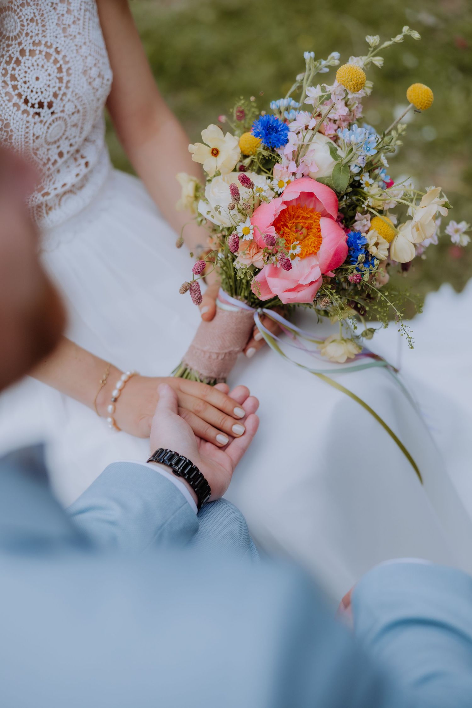 Bride and groom holding hands with a colorful wildflower wedding bouquet featuring pink peonies and blue flowers.