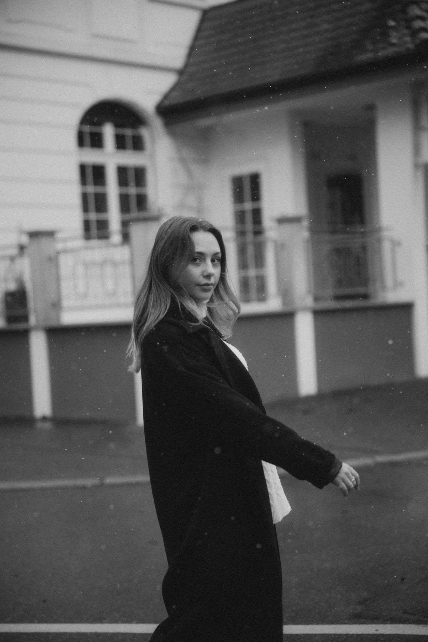 Black and white portrait of someone in a dark coat turning to pose outside a house with arched windows.