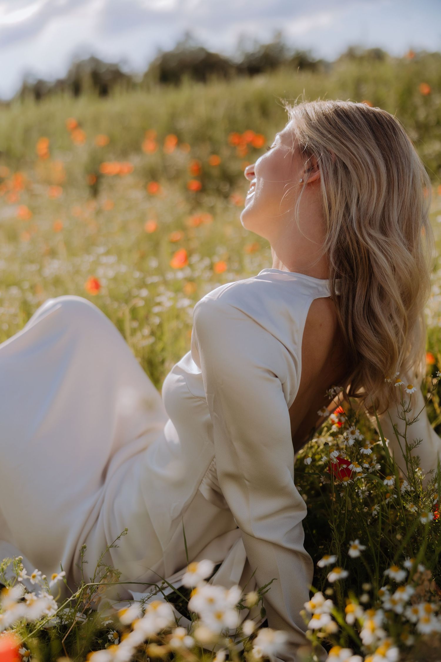 Smiling blonde woman in white dress sitting in a wildflower field with red poppies and daisies on a sunny day.