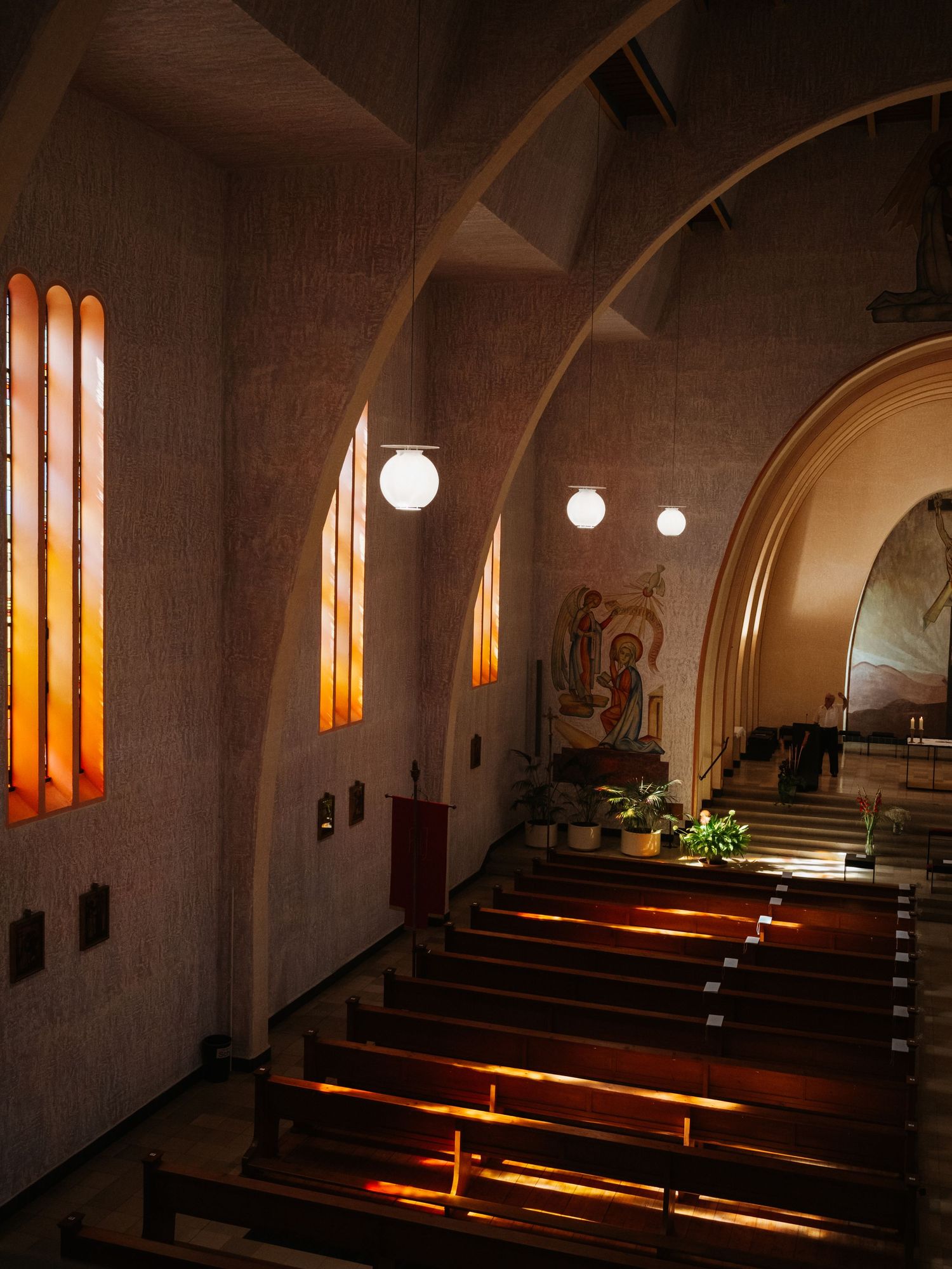 Sunlight streams through narrow stained glass windows in a dimly lit church interior with wooden pews and stone walls.