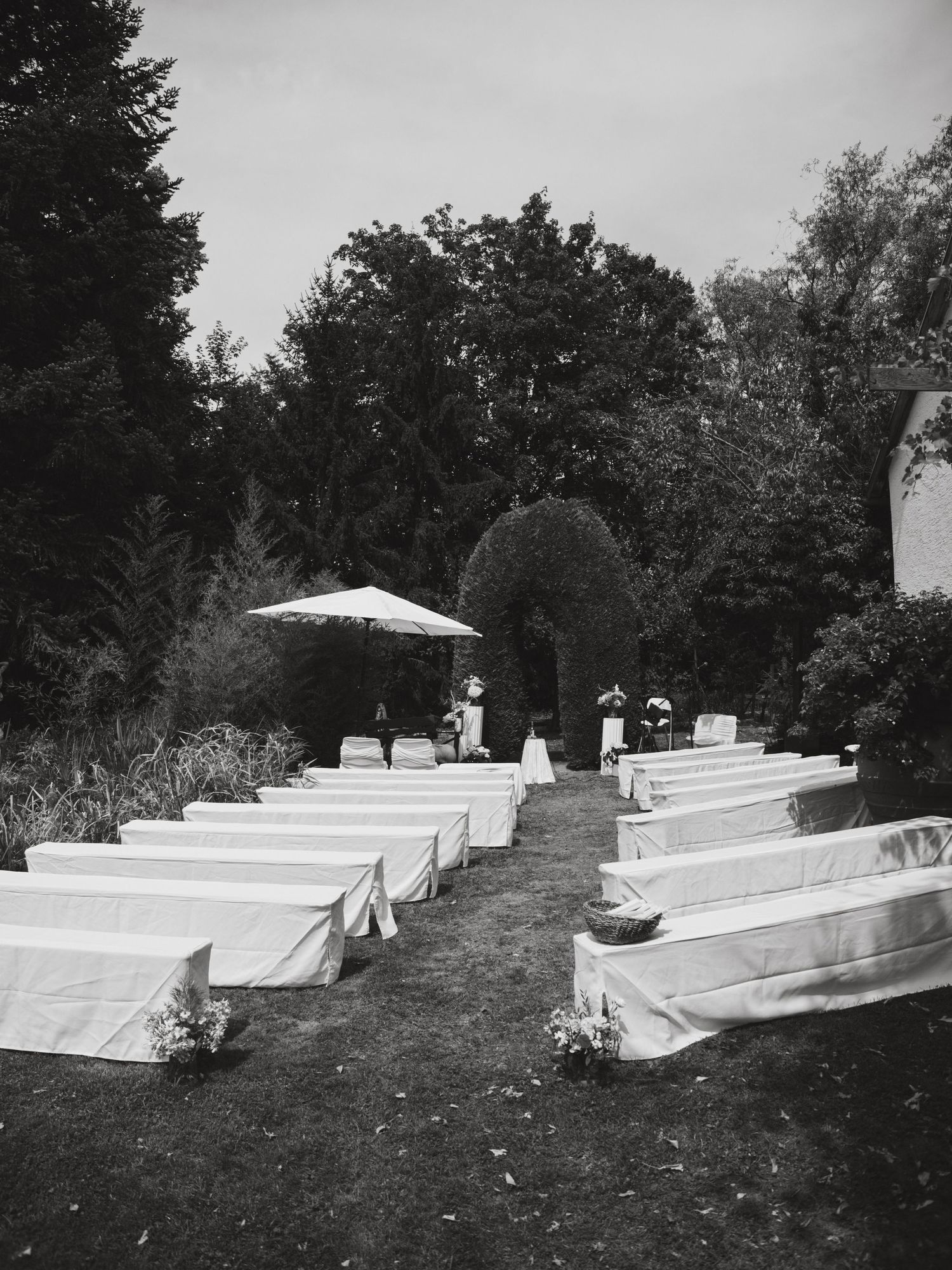 Black and white photo of an outdoor wedding ceremony setup with rows of white-covered chairs and an umbrella at the end.