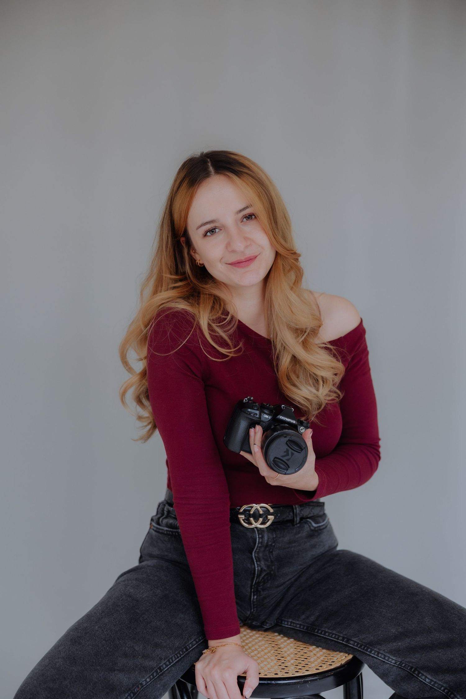 Person in burgundy sweater and dark jeans poses casually while holding a camera against gray background.