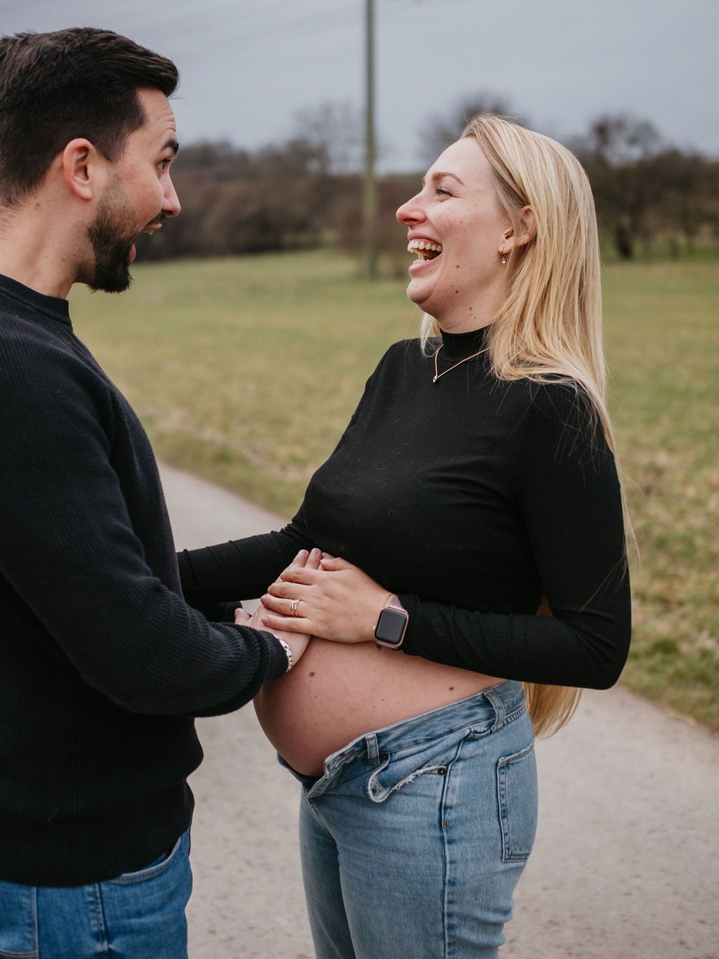 A couple shares a joyful moment on a path in nature, wearing casual black tops and jeans.