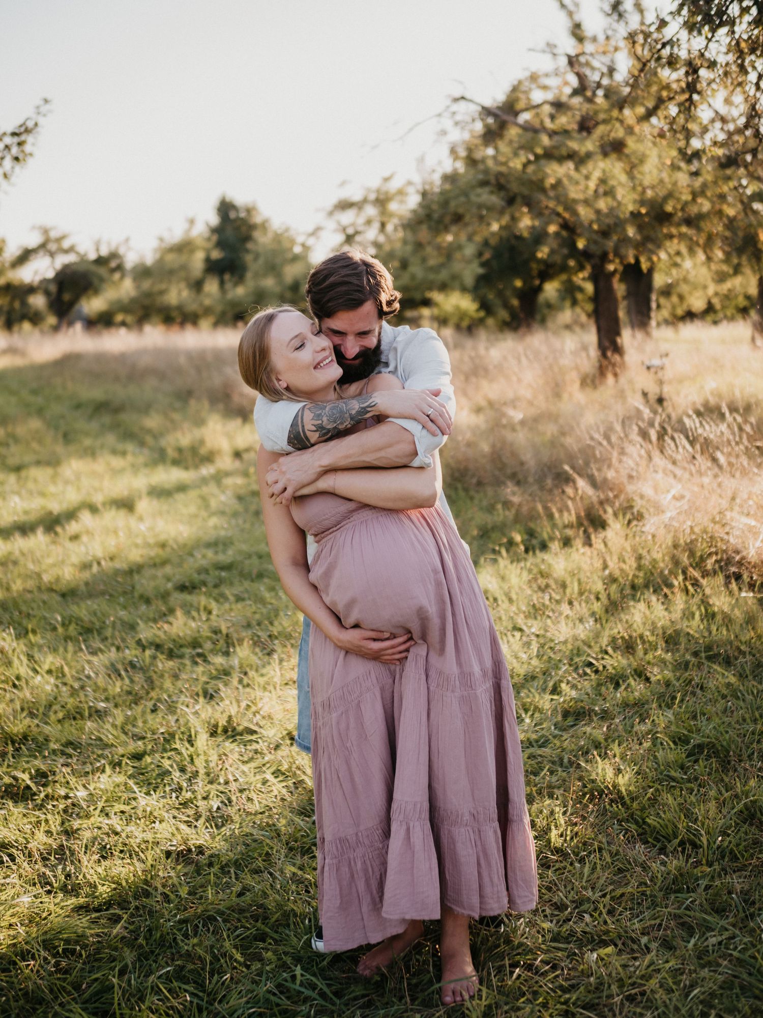 A couple shares tender embraces in a sun-drenched field during golden hour, wearing casual attire.