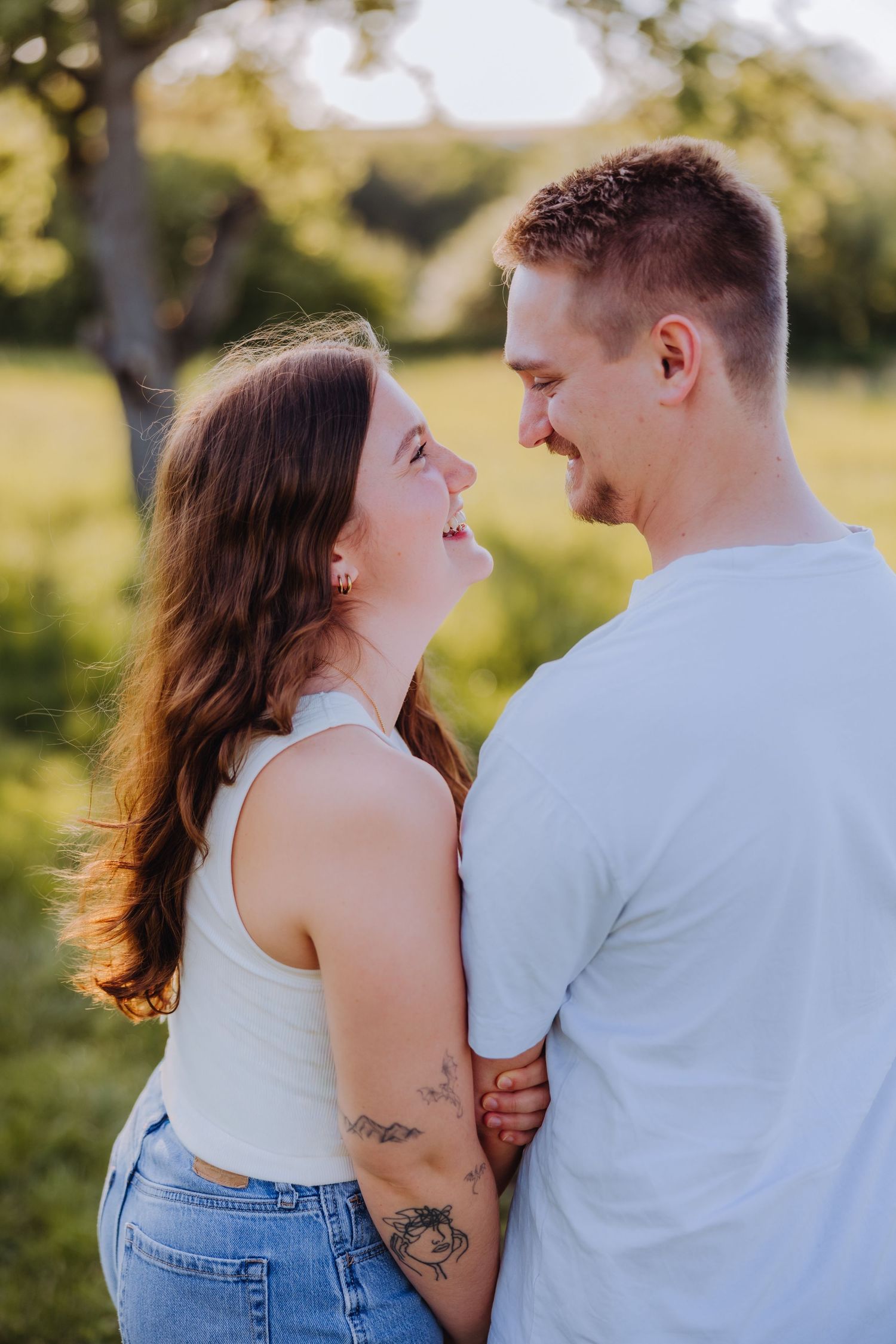 A smiling couple in white tops embraces outdoors, sharing a joyful moment in a sunlit green park setting.
