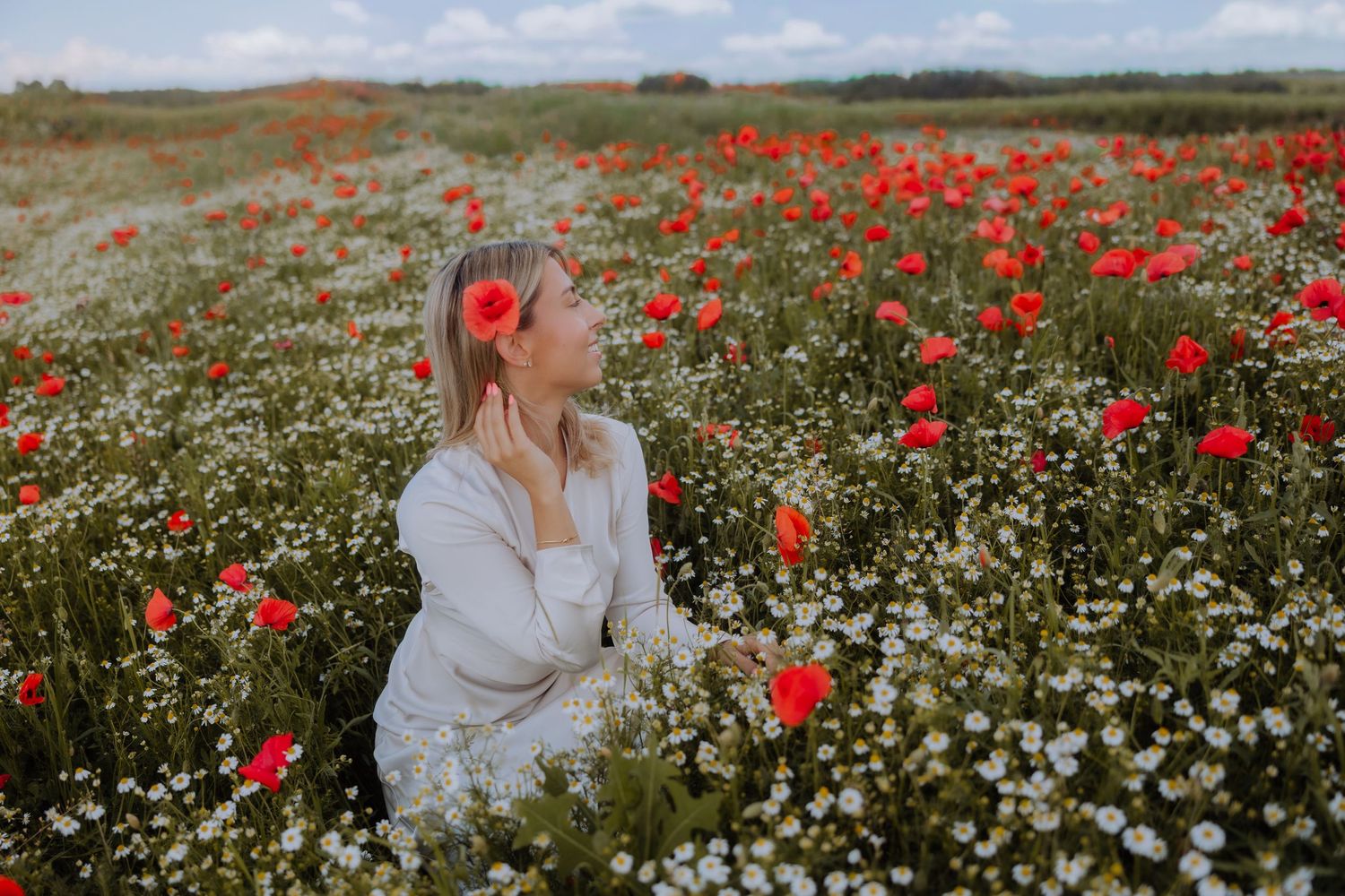 Woman in white dress sitting in a vibrant field of red poppies and white wildflowers on a sunny day.