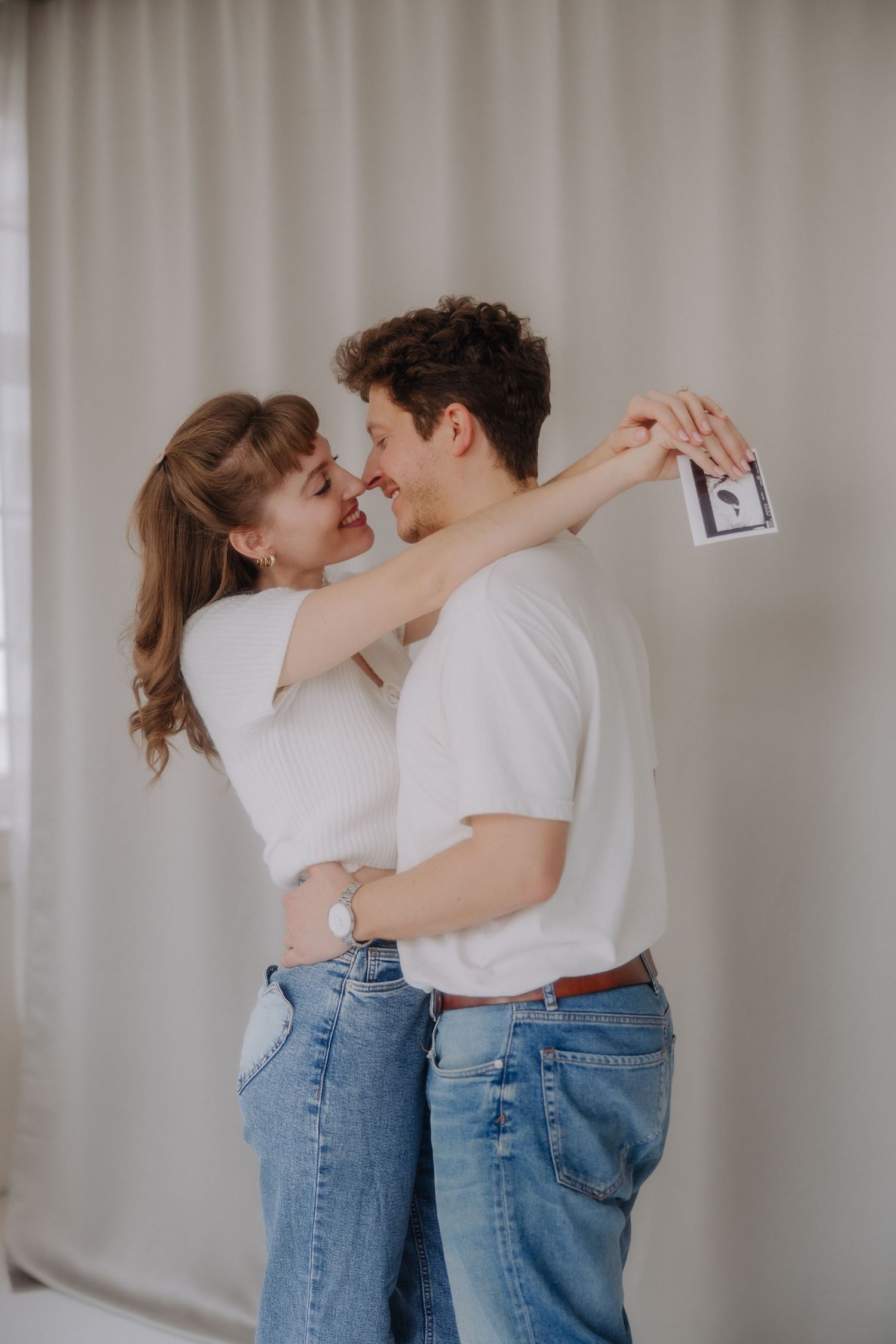 A romantic couple in matching white shirts and blue jeans dance together against a neutral wall.