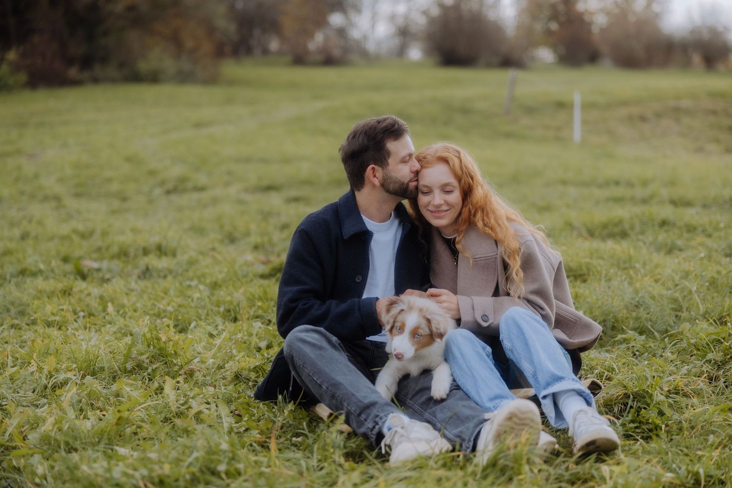 A couple sits together on grass with their golden retriever puppy on a cloudy autumn day.