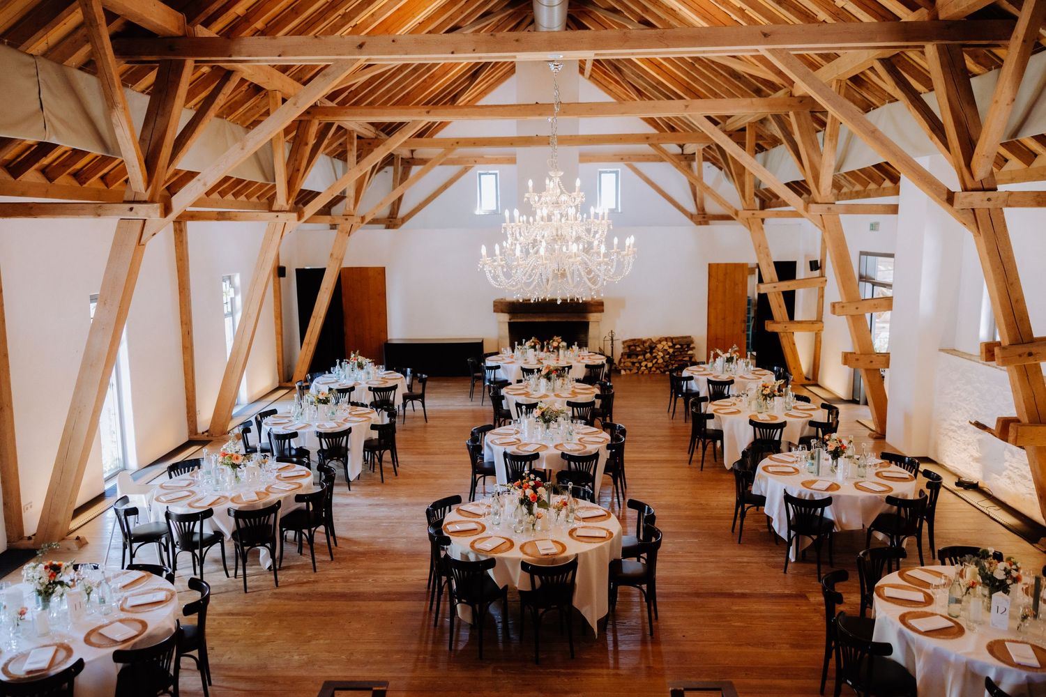 Elegant barn wedding reception with chandelier, round tables, white linens, and wooden beam ceiling.