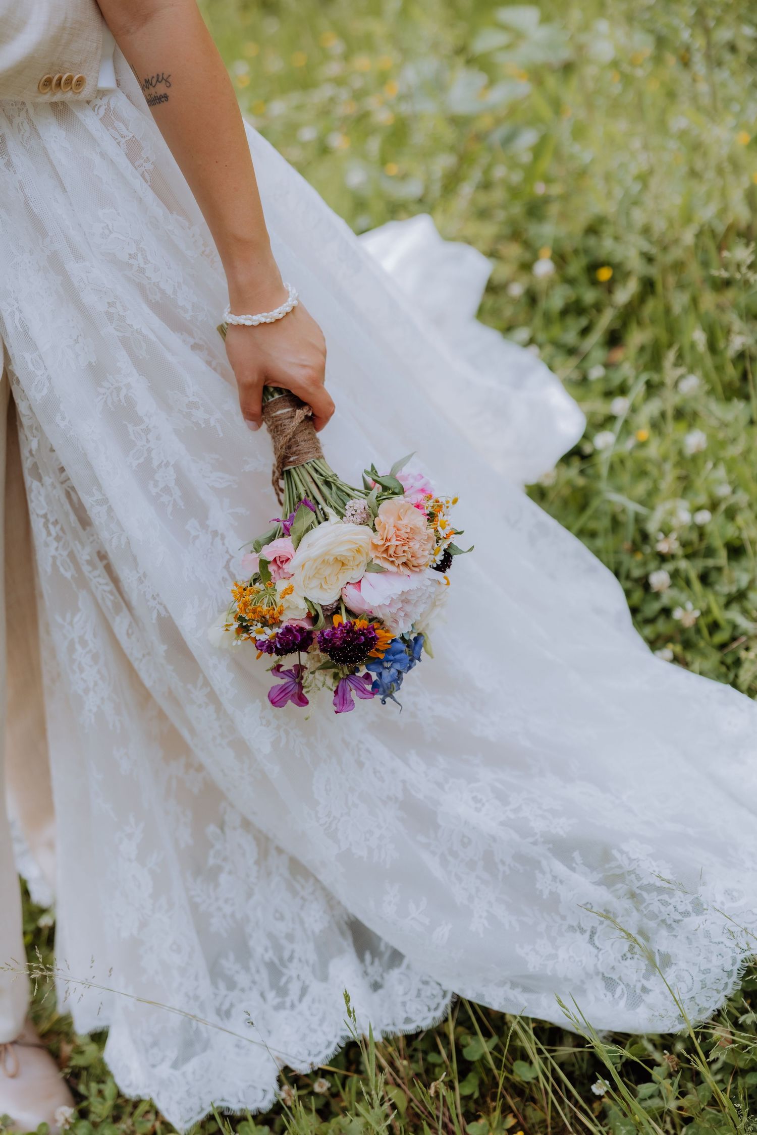Bride in lace wedding gown holding colorful wildflower bouquet with pink, purple, and yellow blooms on green meadow.