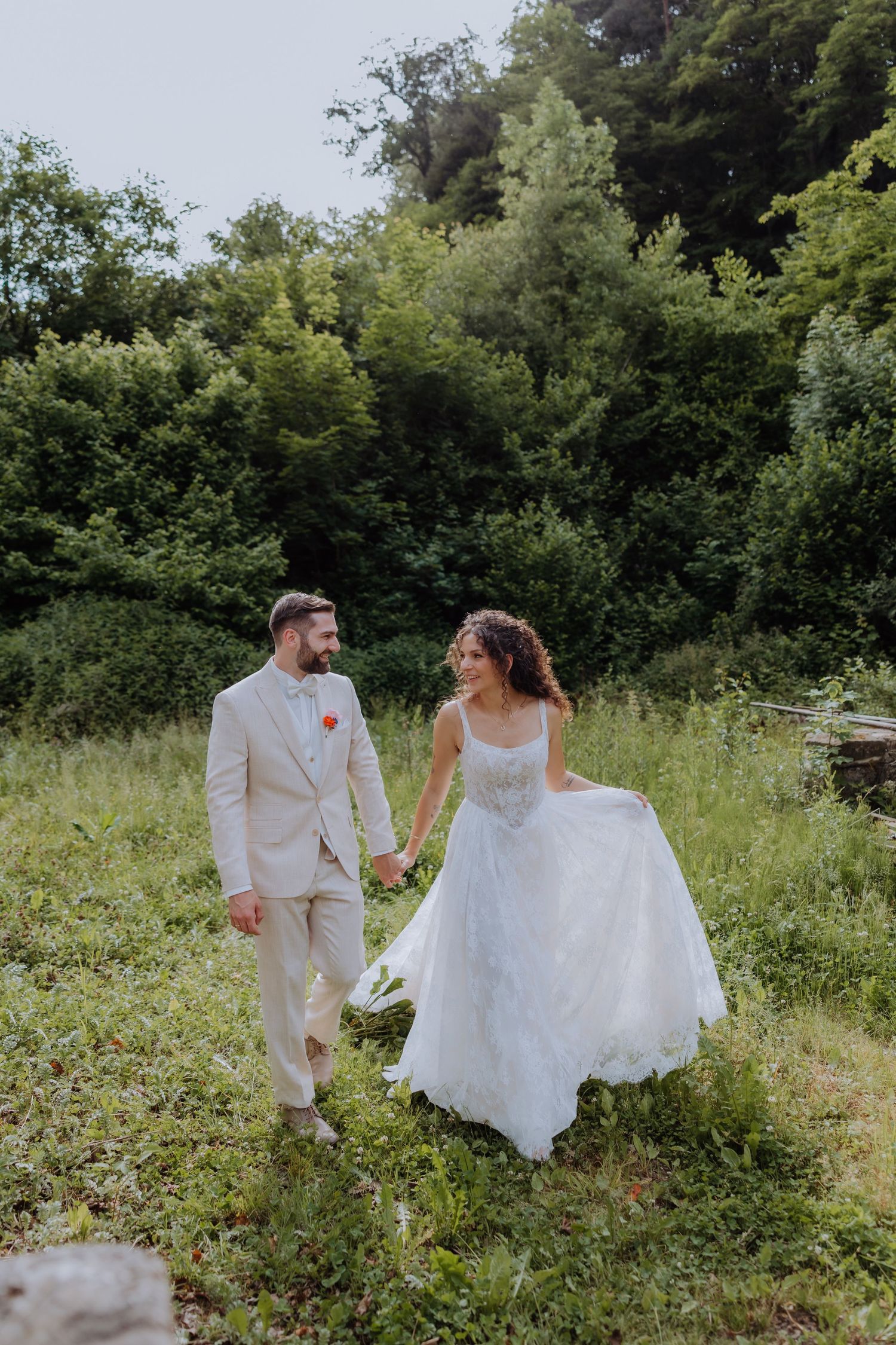 Bride and groom holding hands walking through lush green meadow, bride in white gown, groom in beige suit.
