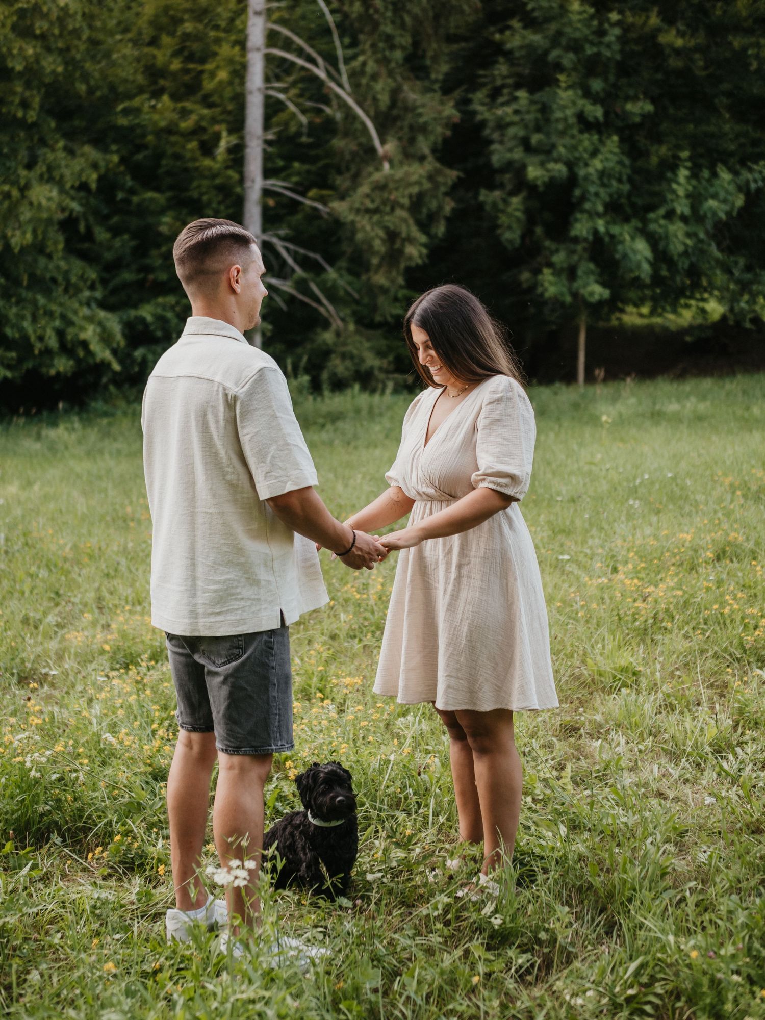 A couple stands together in a grassy meadow on a sunny summer day holding hands in matching white outfits.