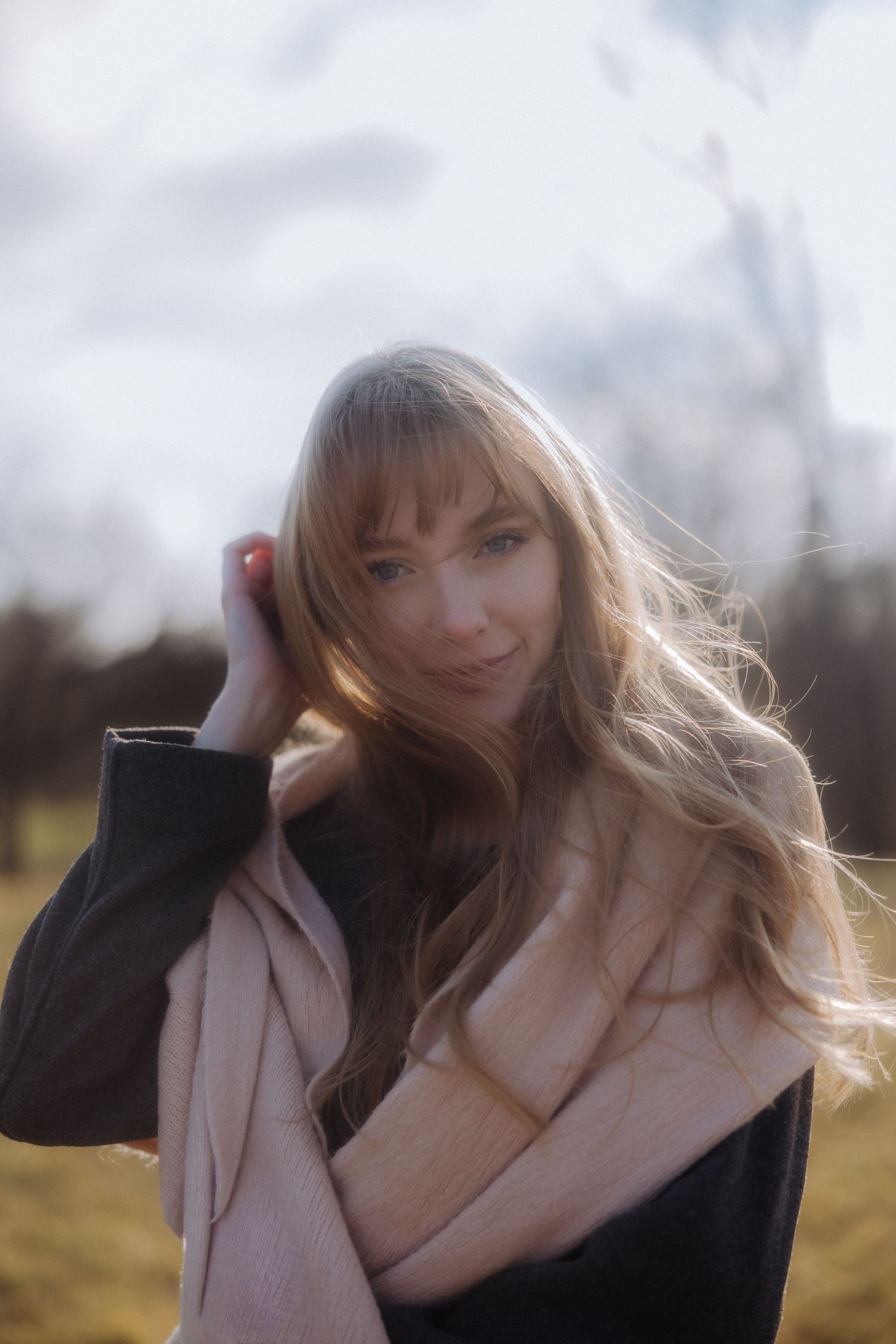 A dreamy outdoor portrait with soft lighting shows a woman in a pink scarf and black sweater with windswept hair.