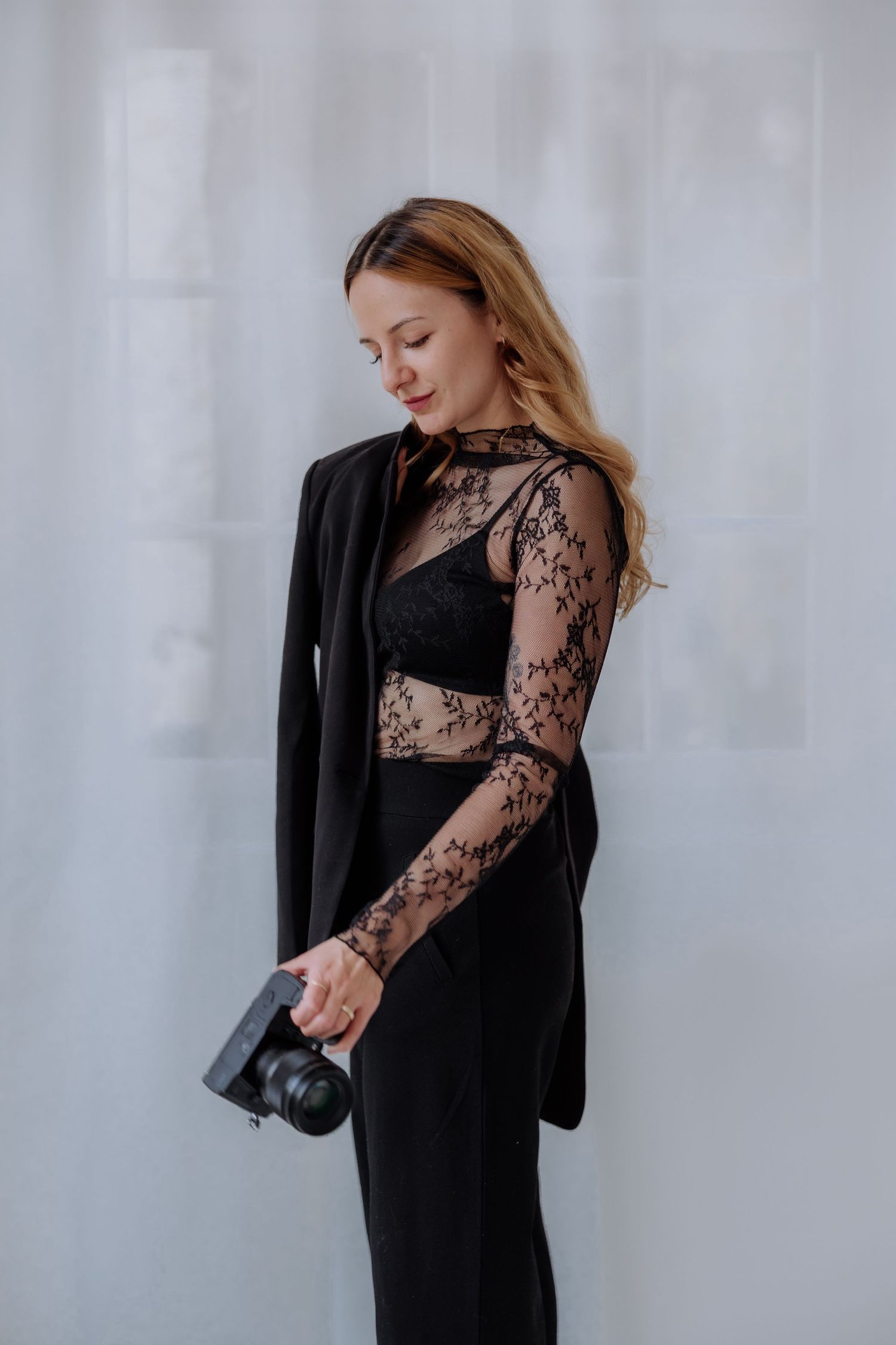A woman in a black lace dress and blazer poses elegantly against a white backdrop in a fashion photoshoot series.