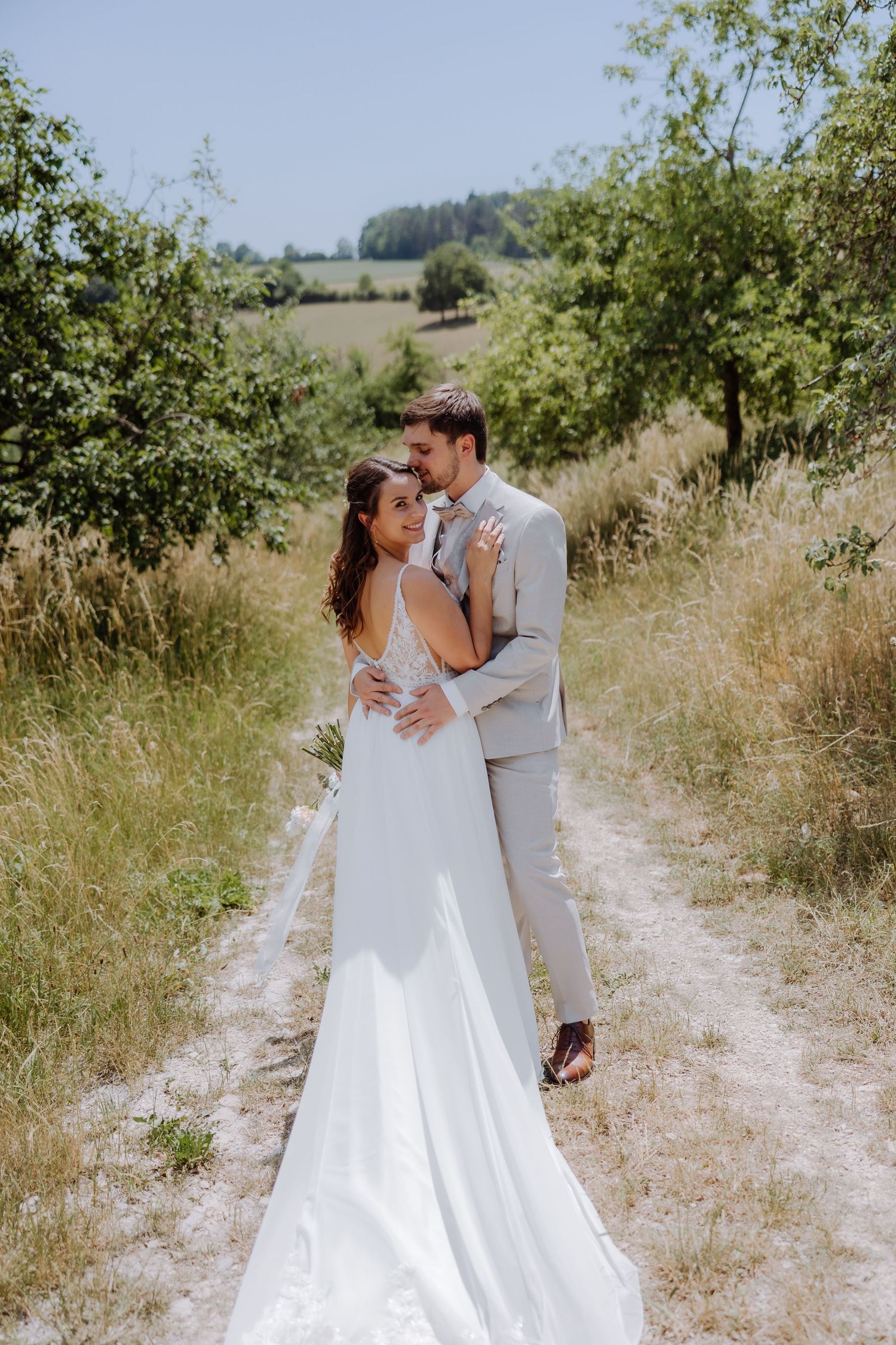 Bride in white gown and groom in gray suit embrace on a scenic countryside path surrounded by greenery.